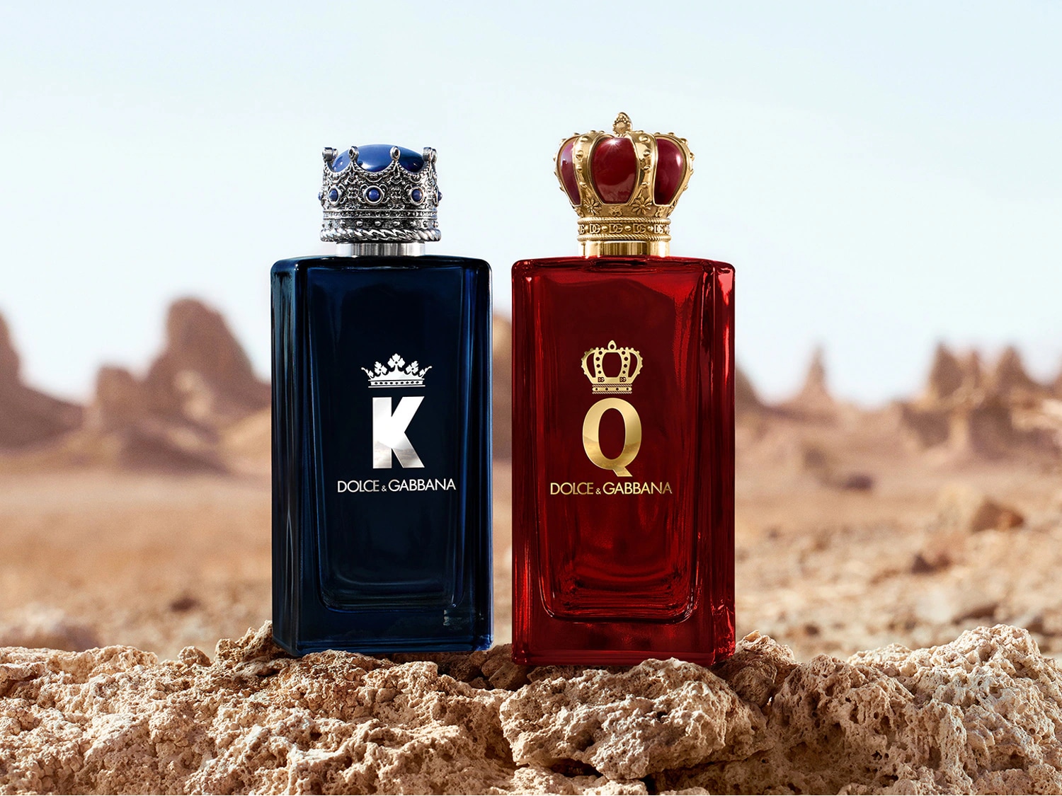 Twee rechthoekige Dolce & Gabbana parfumflessen, een blauwe met de letter K en een rode met de letter Q, beide met een kroon op de dop, staan op een rotsachtige ondergrond.