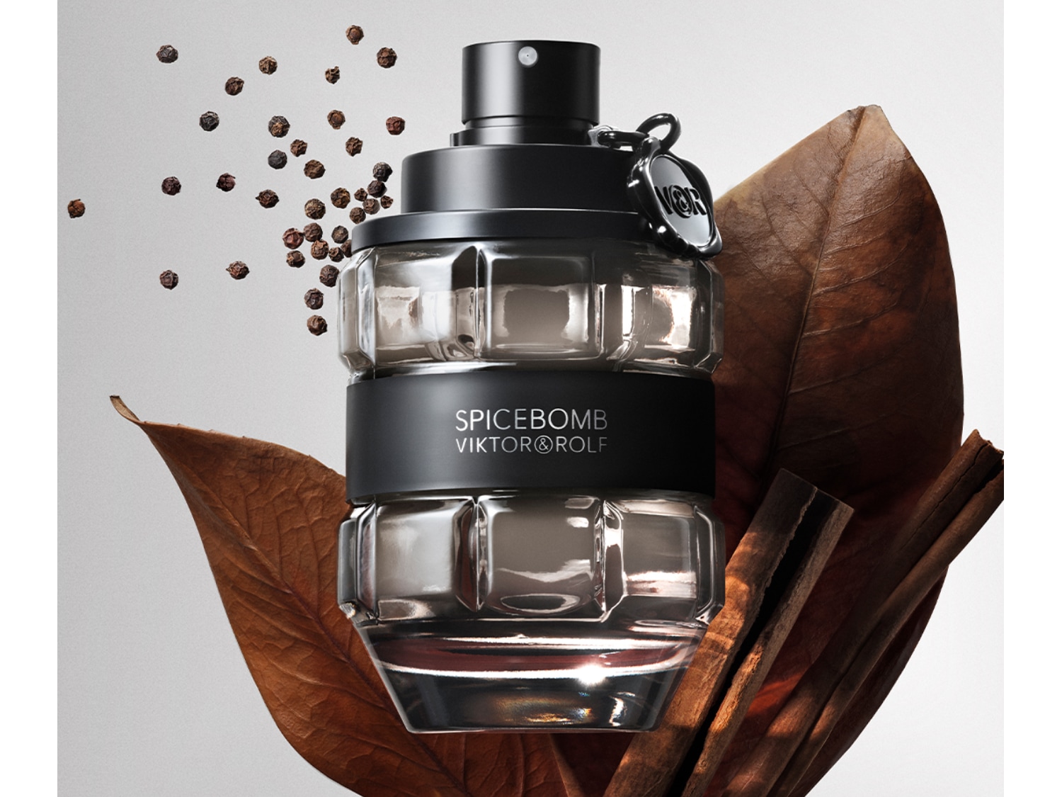 Le parfum Spicebomb de Viktor&Rolf dans un flacon en verre quadrillé avec une bande noire, entouré de feuilles brunes, de bâtons de cannelle et de grains de poivre.