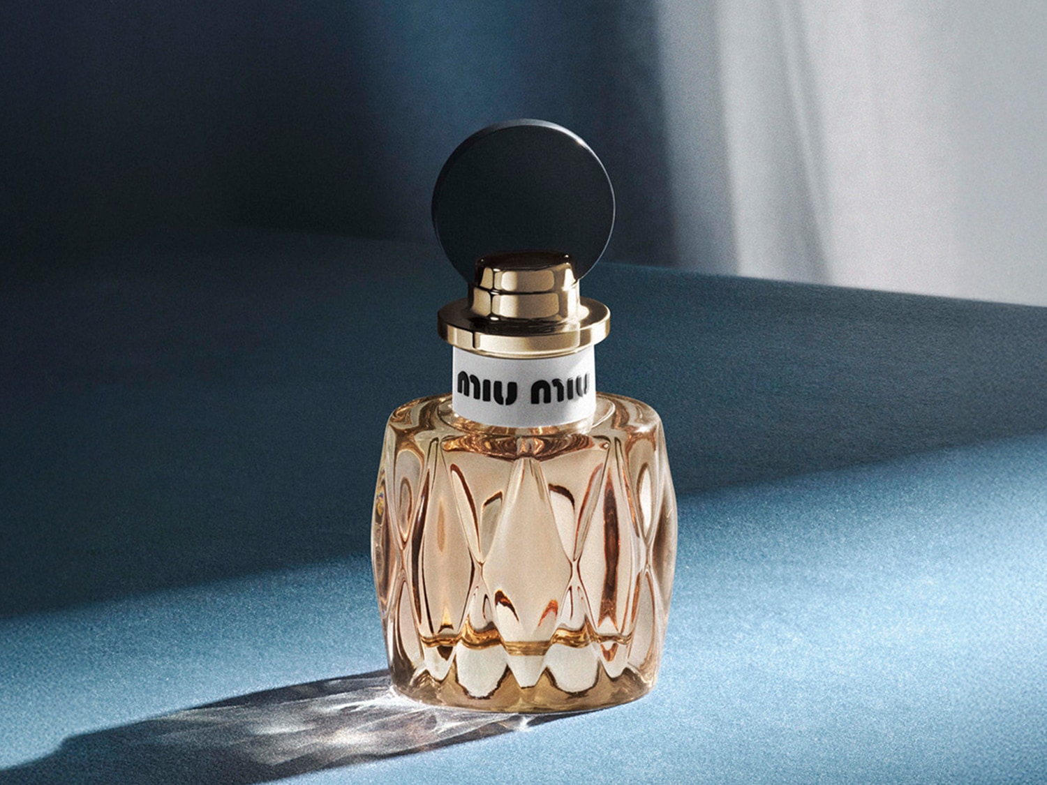 Découvrez le parfum Miu Miu, présenté dans un flacon en verre facetté couleur pêche, surmonté d'un bouchon noir élégant et d'un col doré.