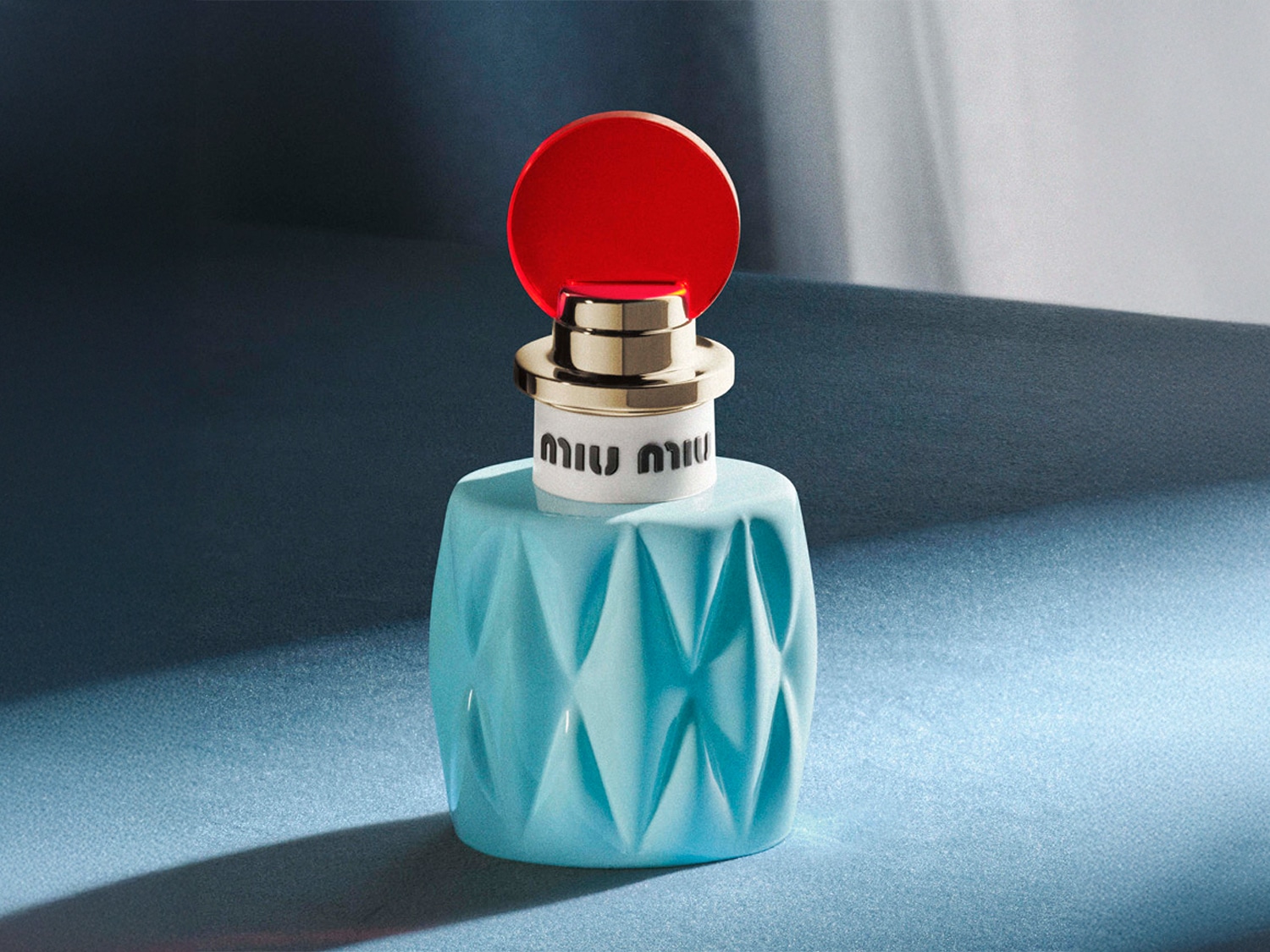 Découvrez le parfum Miu Miu, présenté dans un flacon bleu turquoise aux motifs géométriques, surmonté d'un bouchon rouge vif et doré.