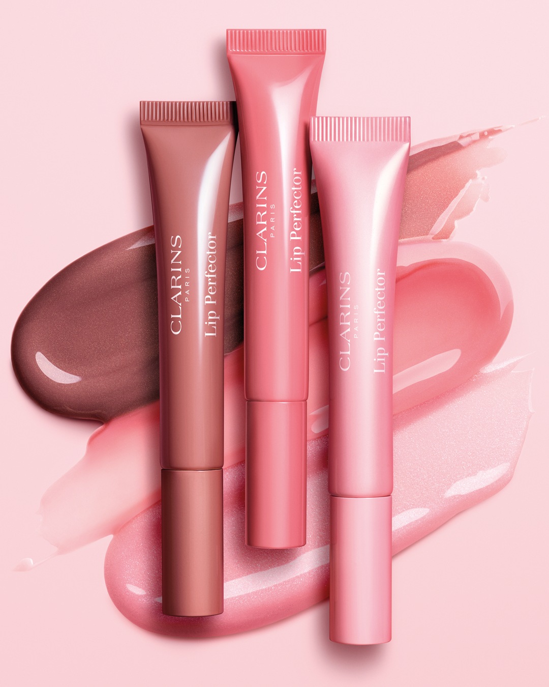 Drie Clarins Lip Perfector tubes in verschillende tinten roze en bruin, liggend op een roze achtergrond met glanzende vegen van de lipgloss.