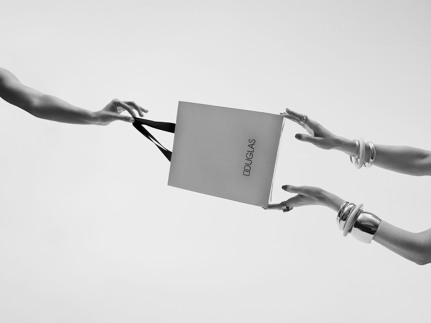 Een Douglas tas wordt doorgegeven tussen twee paar handen, versierd met ringen en zilveren armbanden, tegen een heldere achtergrond.