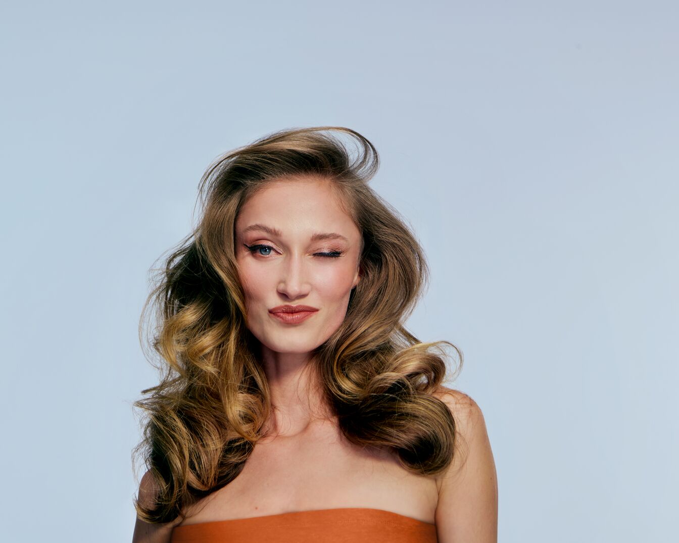 Een vrouw met golvend bruin haar en een oranje top knipoogt speels naar de camera.