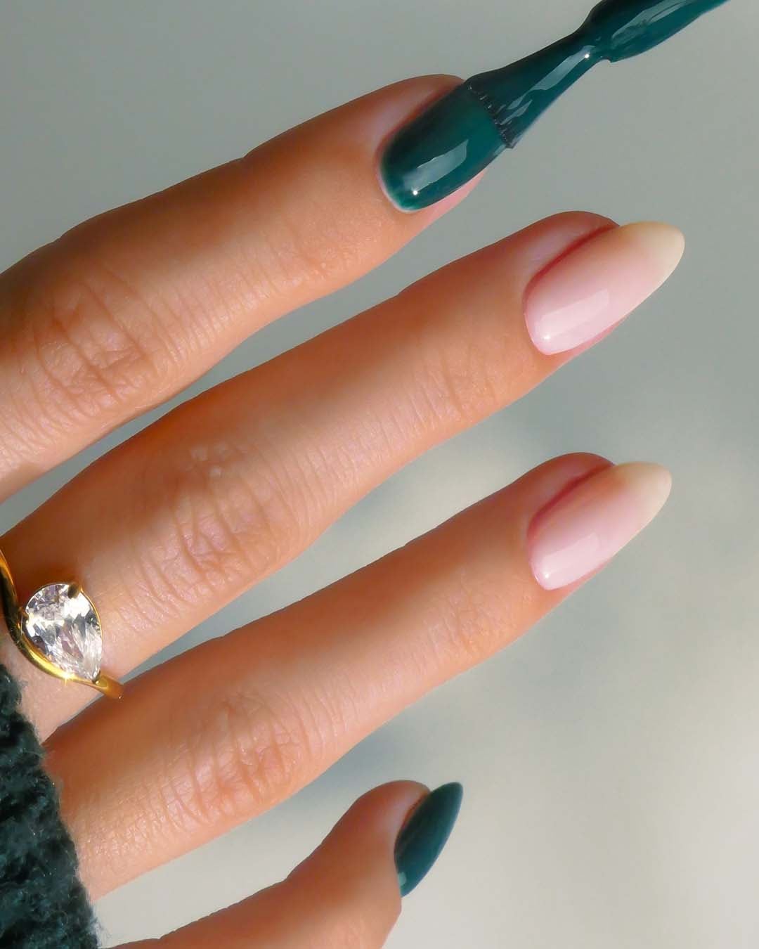 Nahaufnahme einer Hand mit einem goldenen Ring und lackierten Nägeln, wobei ein Nagel dunkelgrün und die anderen hellrosa sind.