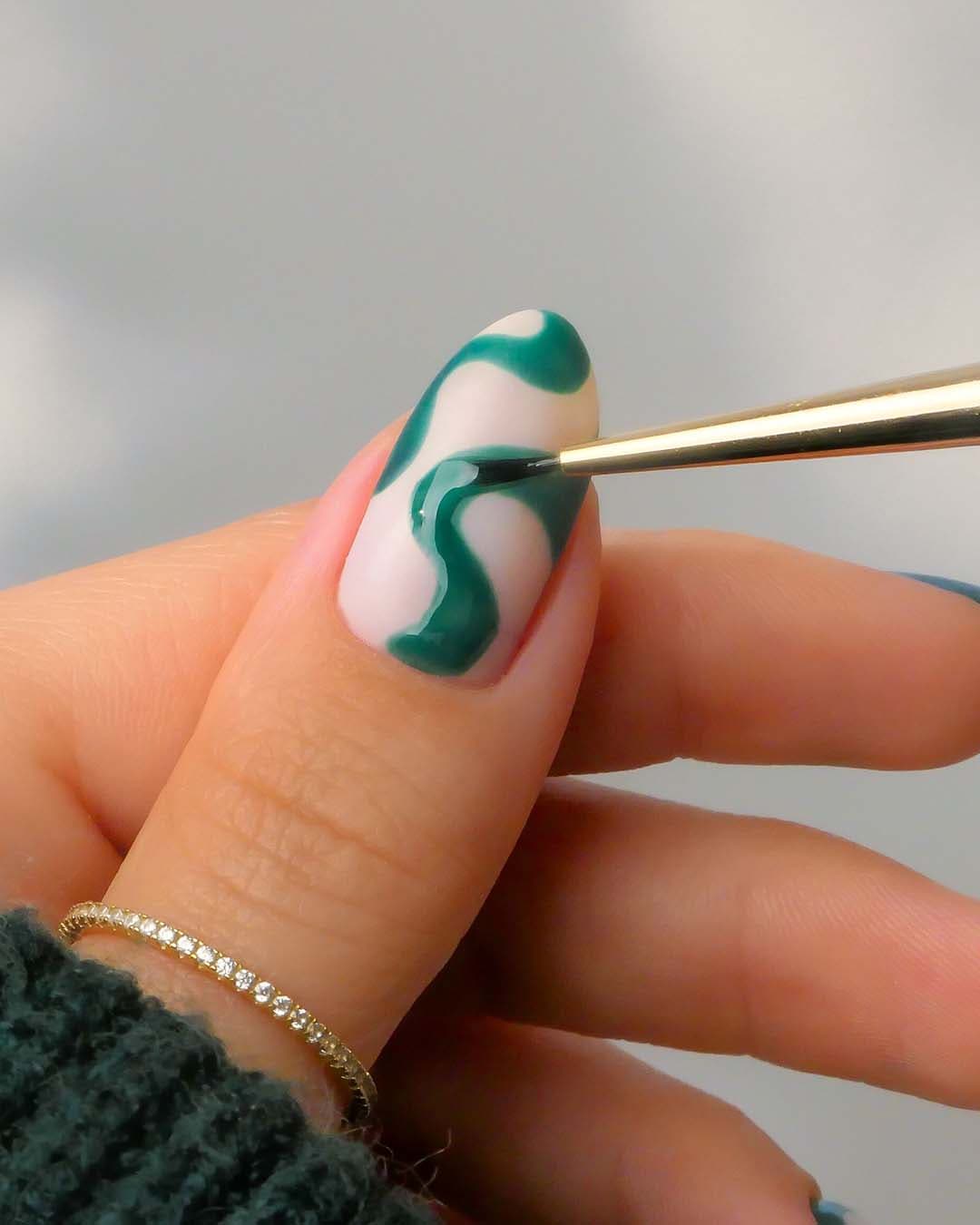 Ein Nagel wird mit einem Pinsel und dunkelgrünem Nagellack in einem geschwungenen Design bemalt.
