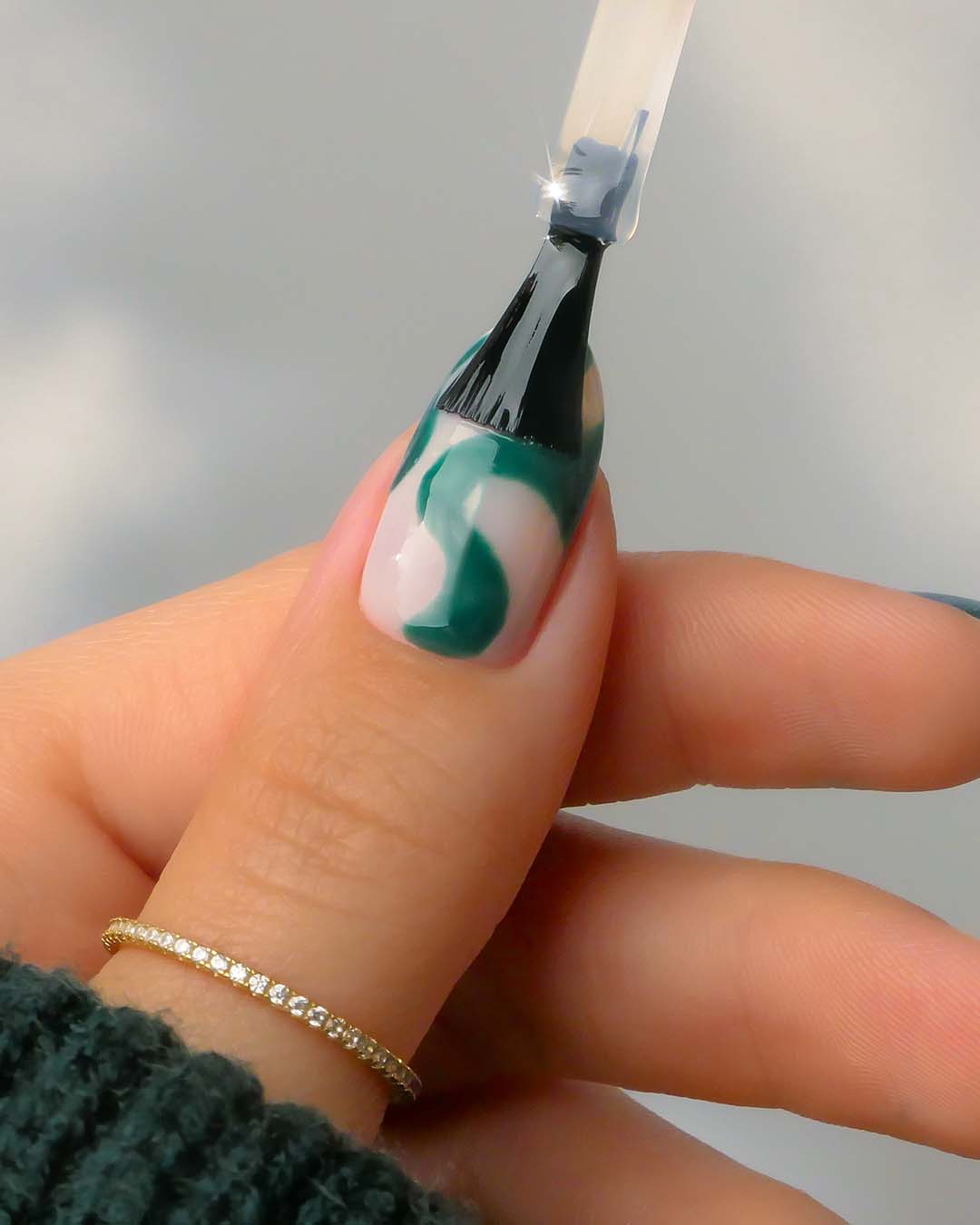 Nahaufnahme einer Hand, die mit einem Pinsel grünen Nagellack in einem Wirbelmuster auf einen Fingernagel aufträgt, der mit einem goldenen Diamantring verziert ist.