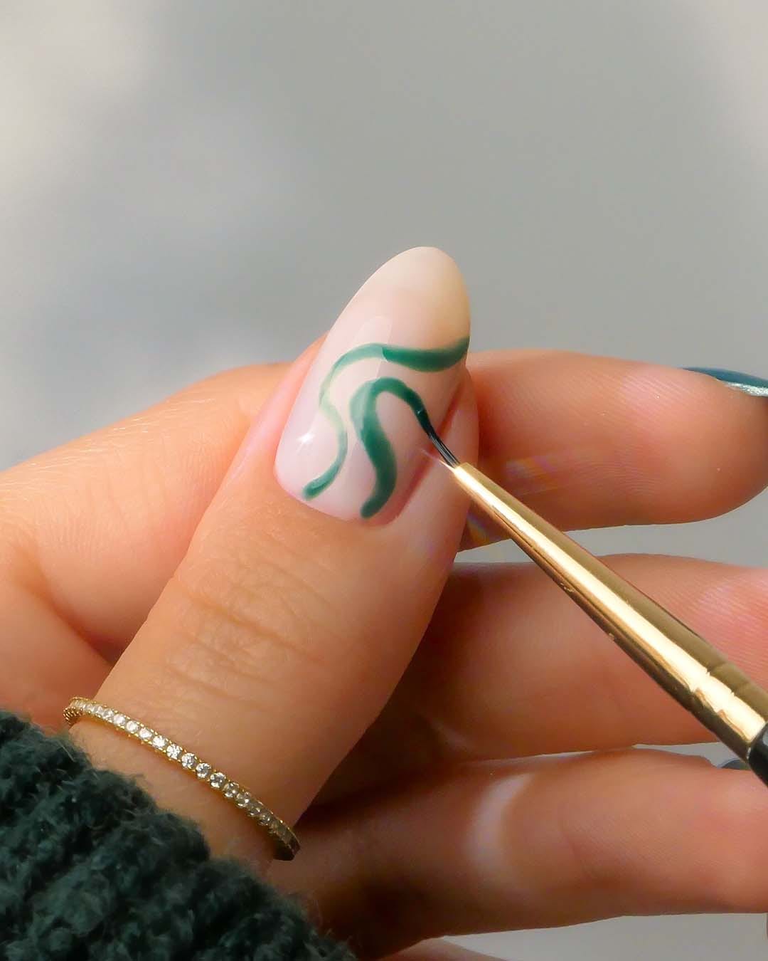 Ein Nagel wird mit einem feinen Pinsel und grüner Farbe mit einem geschwungenen Muster verziert.
