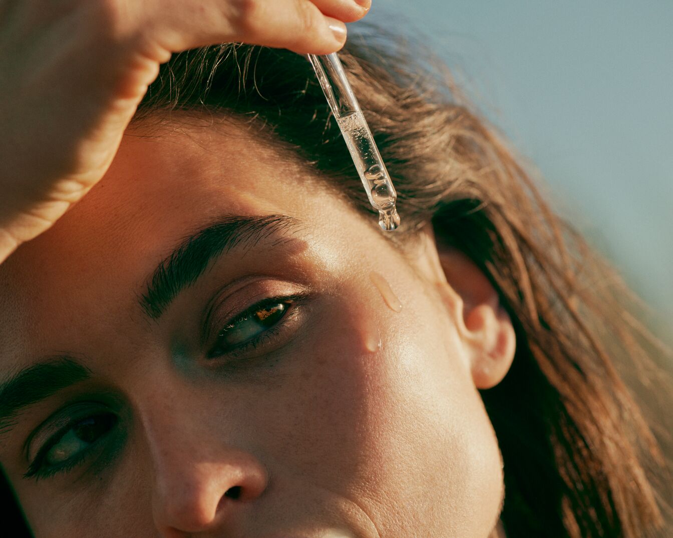 Gros plan sur une femme appliquant un sérum transparent sur son visage à l'aide d'une pipette en verre, pour une peau éclatante et hydratée.