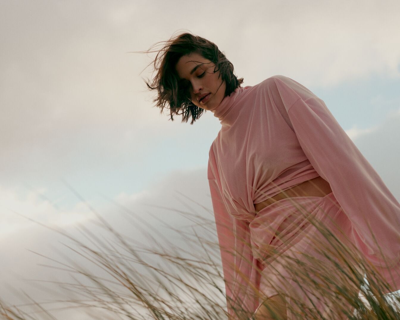 Une femme aux cheveux bruns, vêtue d'une robe rose, se tient dans un champ d'herbe, avec un ciel nuageux en arrière-plan.