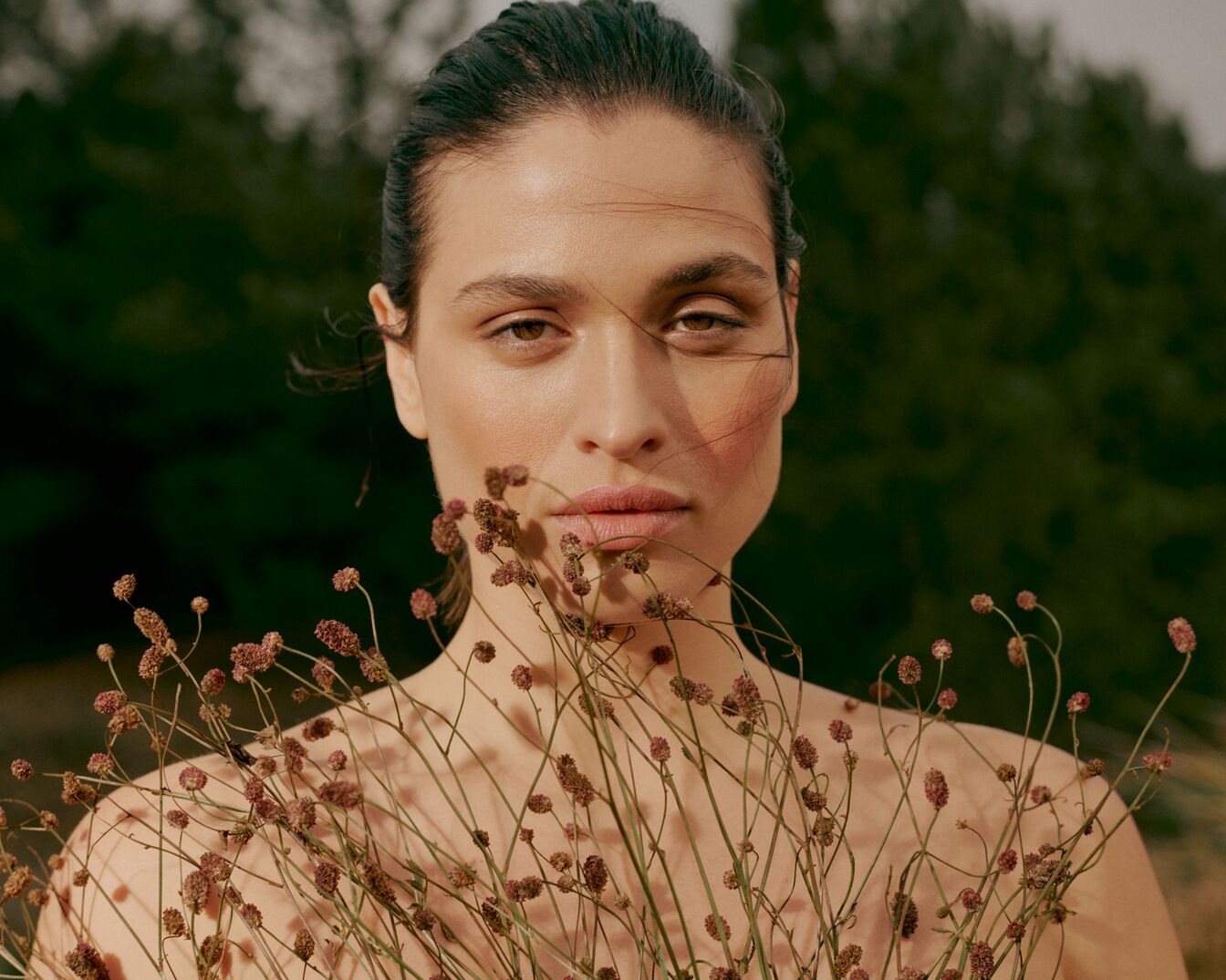 Portrait d'une femme aux cheveux bruns tirés en arrière, tenant un bouquet de fleurs séchées devant un fond de verdure.