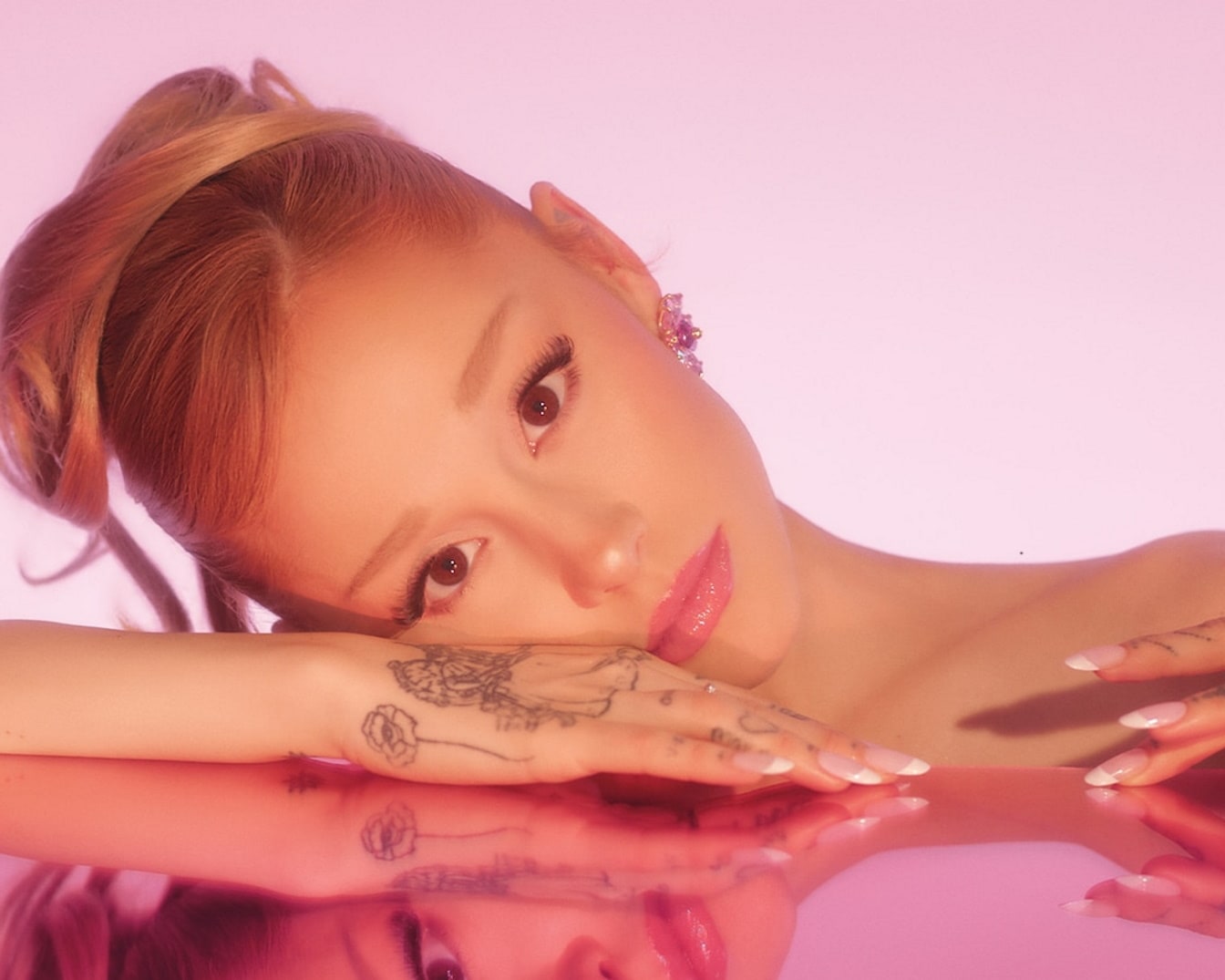 Gros plan d'une femme aux cheveux roux coiffés en queue de cheval haute, la peau claire et un tatouage floral sur la main, reposant sur une surface rose réfléchissante.