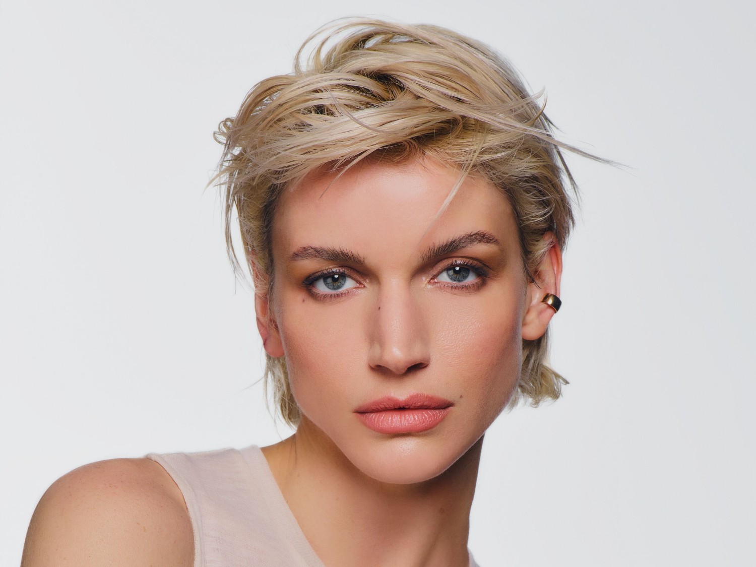 Porträt einer Frau mit blonden, gewellten, kurzen Haaren, hellblauen Augen und einem goldenen Ohrclip vor einem hellen Hintergrund.