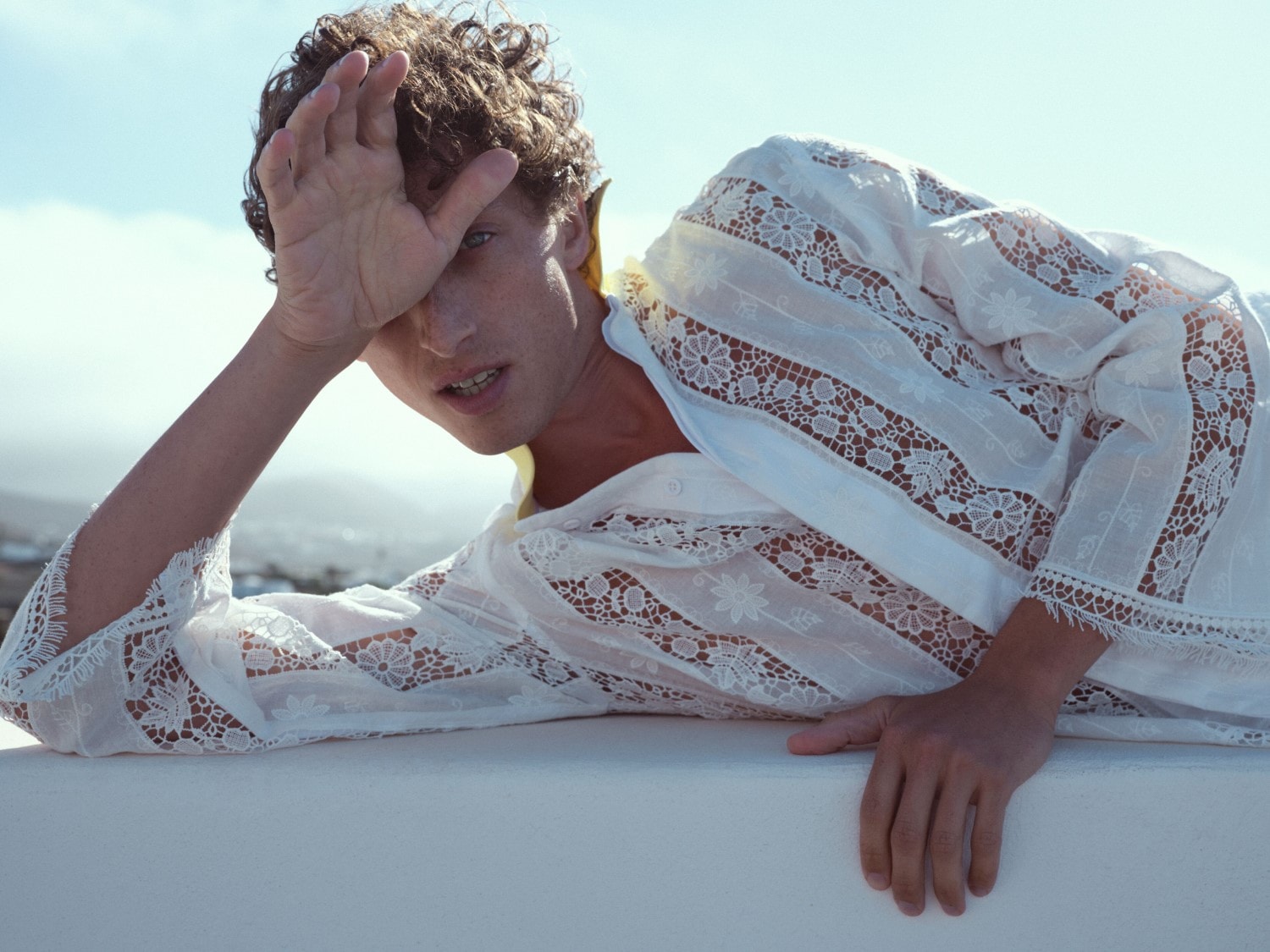 Ein Mann mit lockigem Haar liegt auf einer weißen Oberfläche und trägt ein weißes, durchsichtiges Hemd mit Blumenmuster, während er seine Hand über seine Augen hält und so einen Teil seines Gesichts verdeckt.