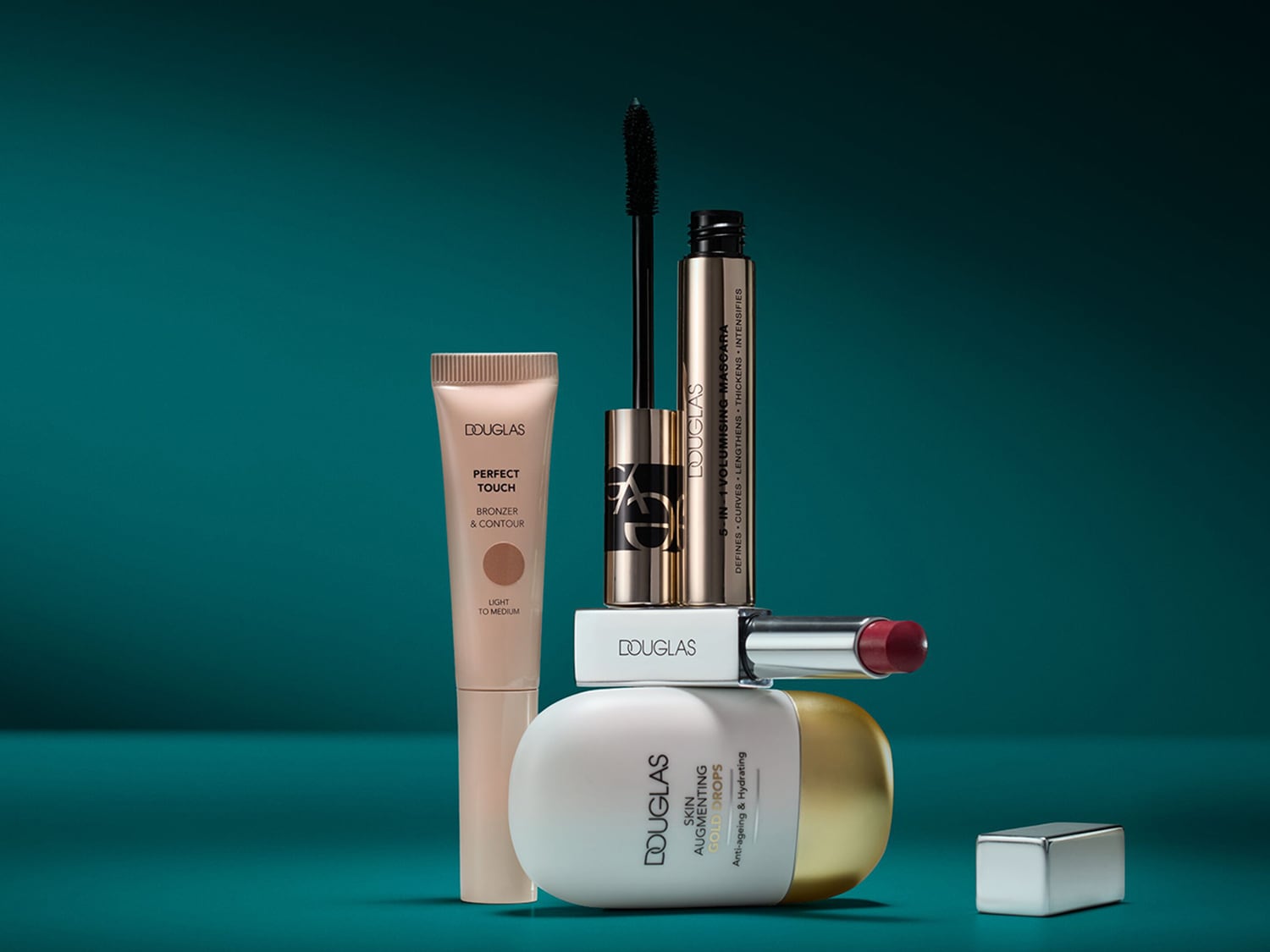 Douglas make-up collectie met Perfect Touch bronzer, 5-in-1 volumising mascara, Skin Augmenting Gold Drops en een rode lippenstift tegen een blauwe achtergrond.