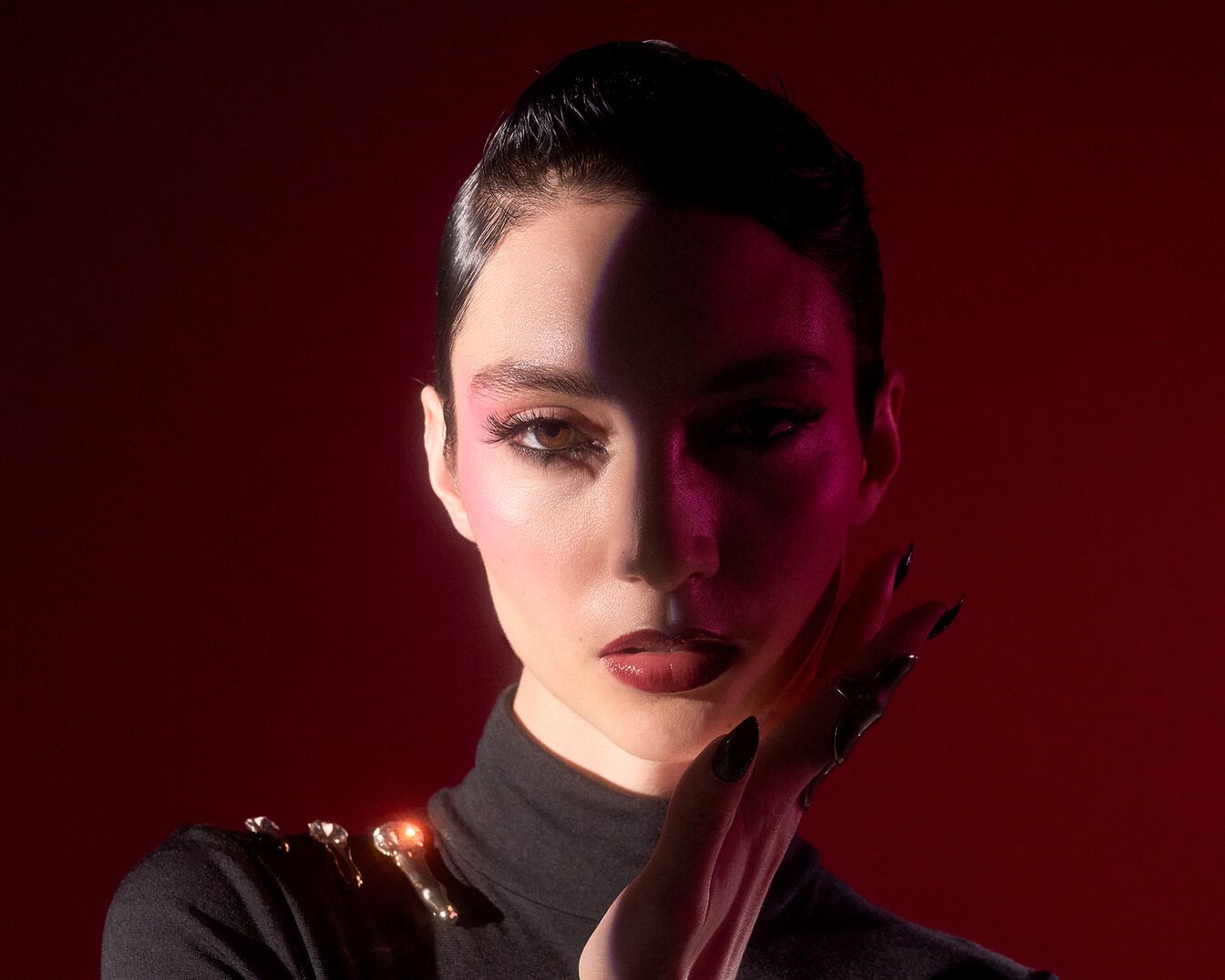 Portret van een vrouw met een strak kapsel, opvallende make-up en lange, zwarte nagels tegen een dieprode achtergrond.