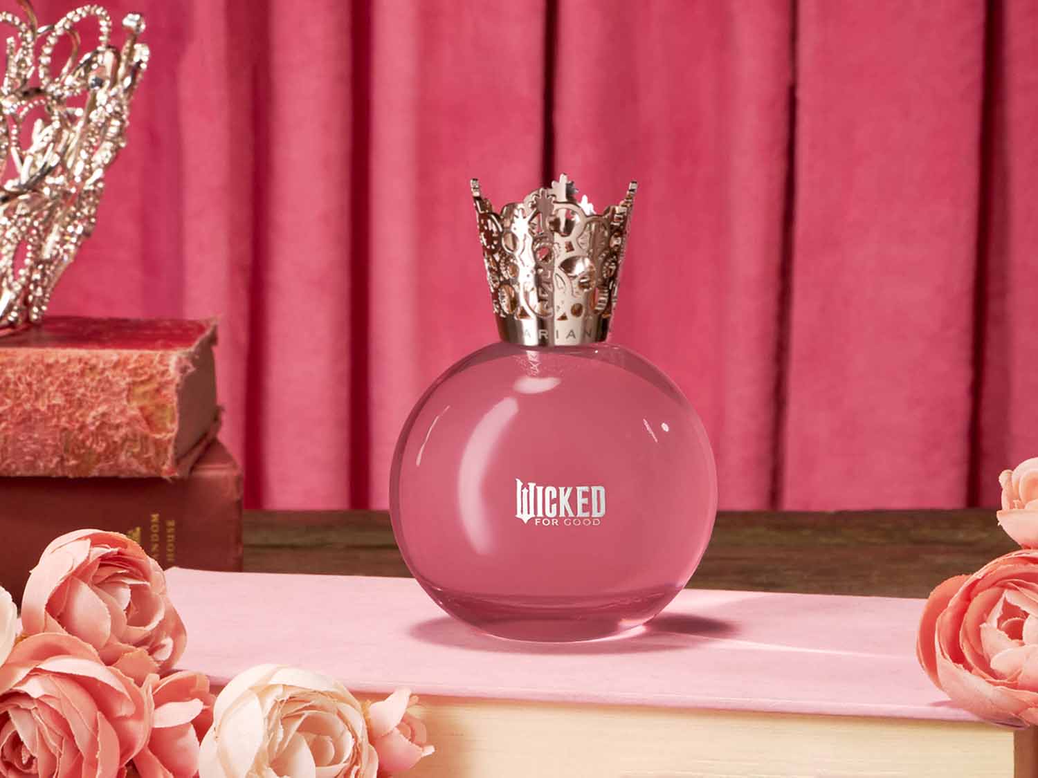 Różowy flakon perfum „Wicked For Good” w kształcie kuli z srebrną nakrętką w kształcie korony, otoczony różami i książkami na tle różowej zasłony.