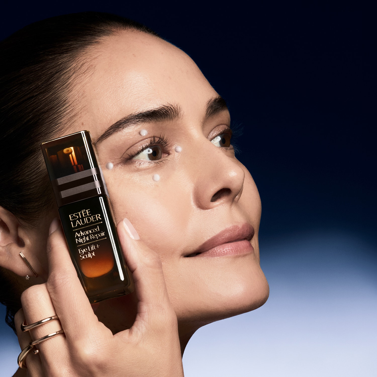 Eine Frau hält eine braune Flasche Estée Lauder Advanced Night Repair Eye Lift + Sculpt Augenserum, während kleine Serum Punkte um ihr Auge aufgetragen sind.