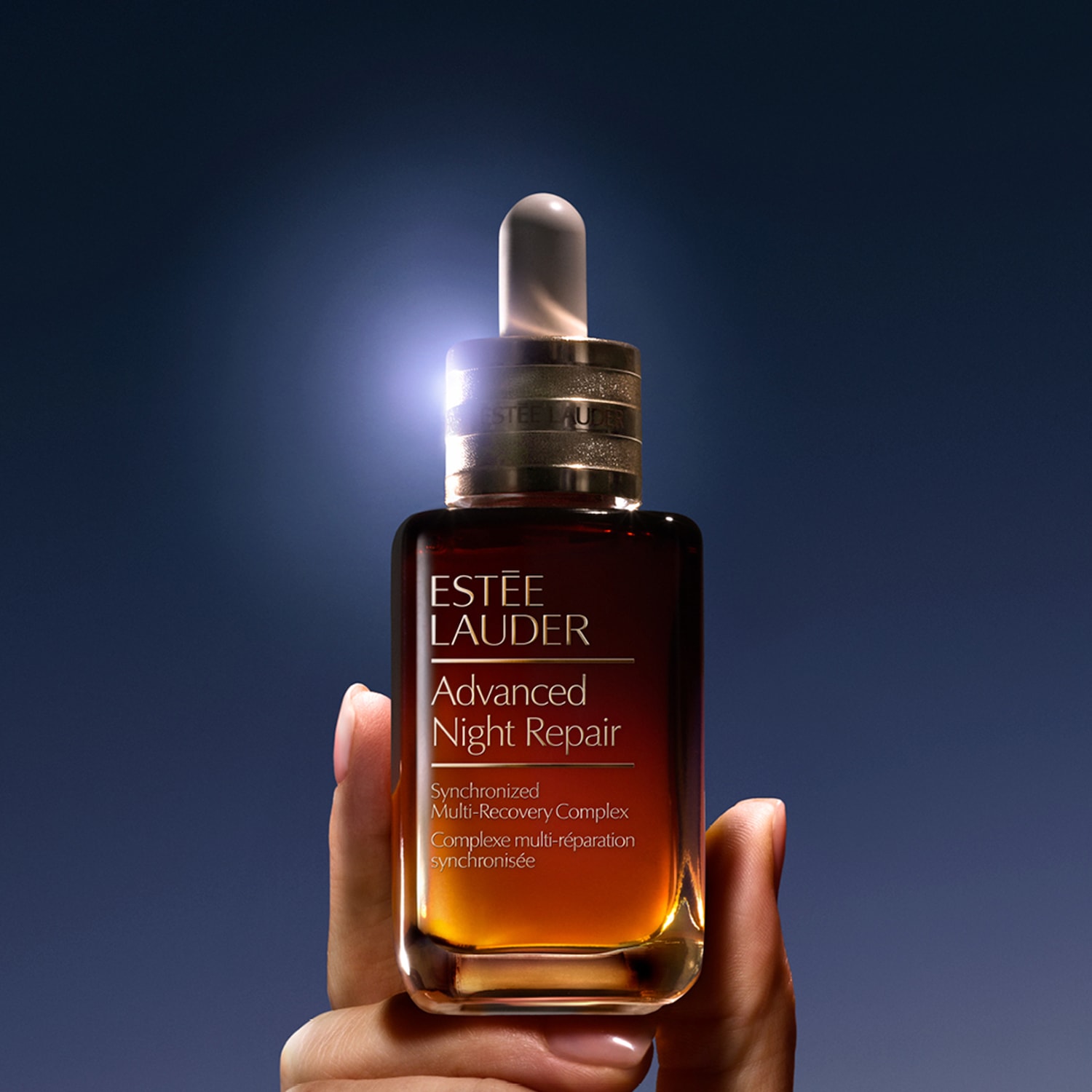 Eine Hand hält eine Flasche Estée Lauder Advanced Night Repair Serum mit Pipette vor blauem Hintergrund.