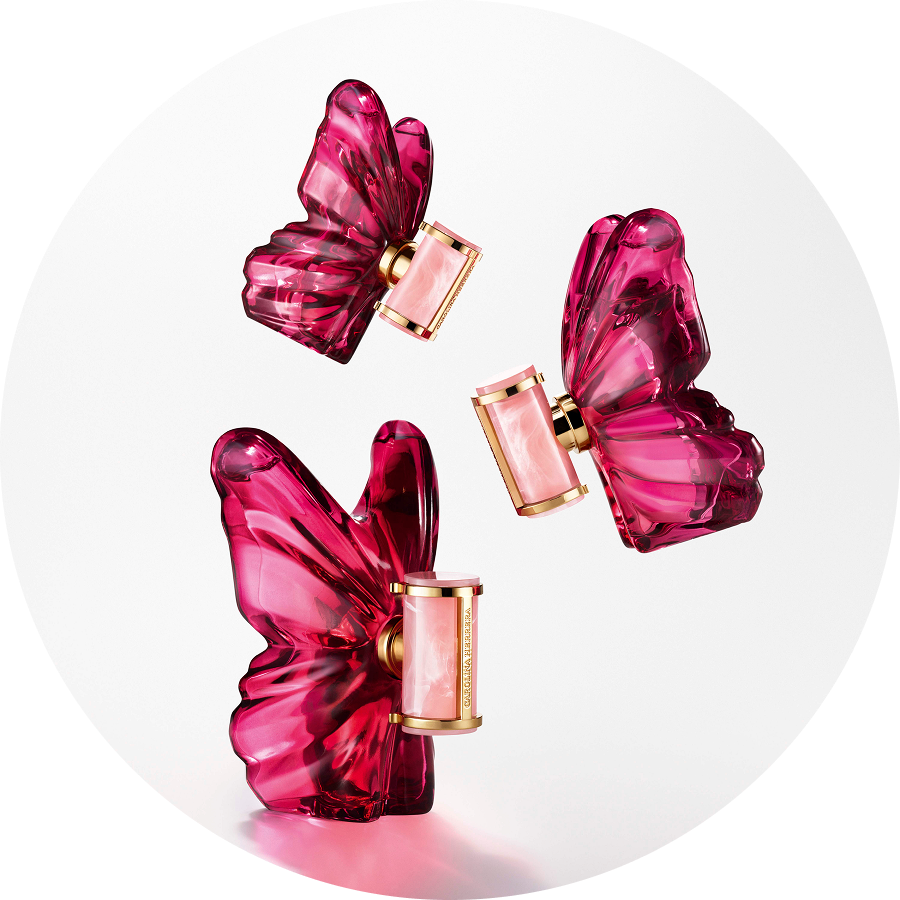 Trzy flakony perfum Carolina Herrera w kształcie różowych motyli z cylindrycznymi, złotymi elementami, unoszące się na białym tle.