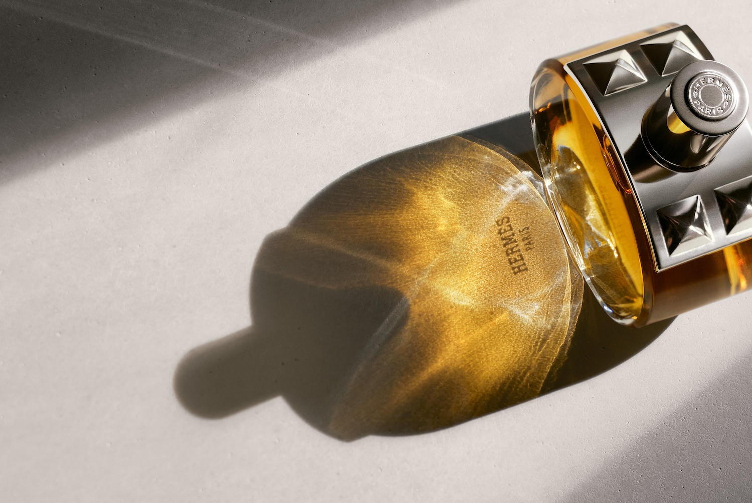 Nahaufnahme eines rechteckigen Flakons von Hermès Paris mit silberfarbenem Verschluss und goldenem Inhalt, der einen warmen Schatten auf eine helle Oberfläche wirft.