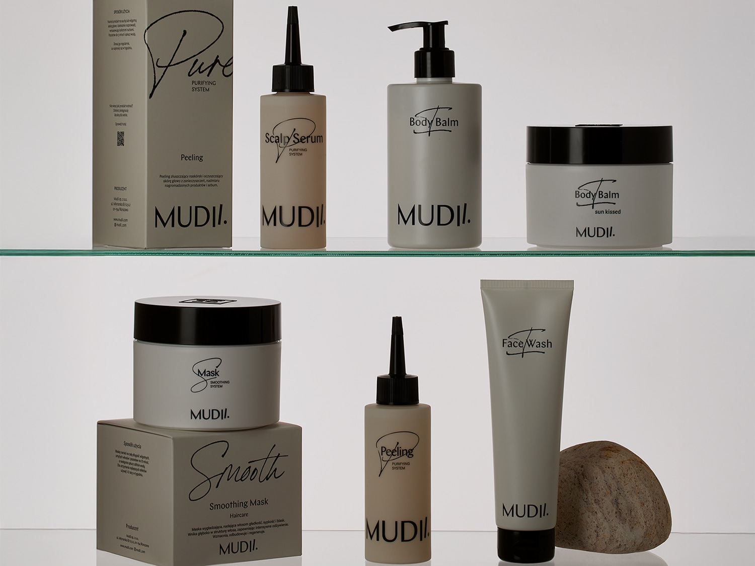 Na szklanej półce stoi seria kosmetyków MUDII w odcieniach szarości, w tym peeling do skóry głowy, balsam do ciała i maska wygładzająca do włosów.