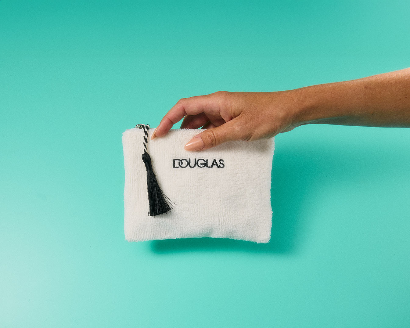 Eine Hand hält eine weiße Kosmetiktasche mit dem Douglas-Logo und einem schwarzen Anhänger vor einem türkisfarbenen Hintergrund.