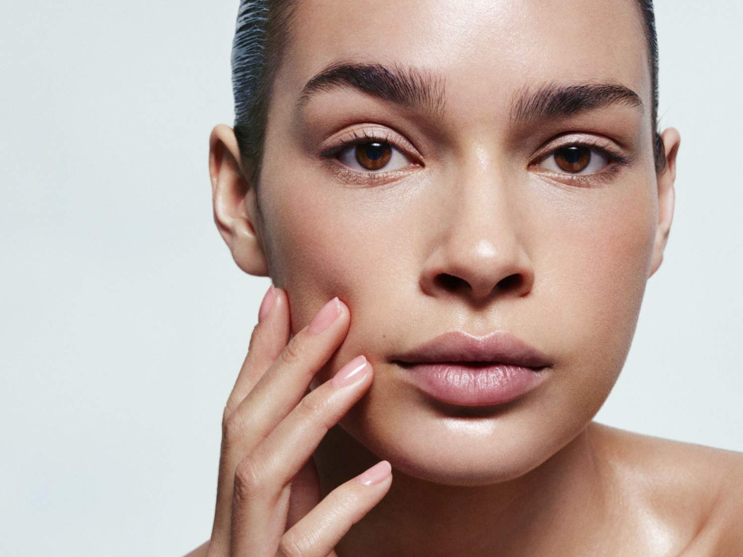 Gros plan sur le visage d'une femme à la peau lisse et éclatante, avec une main délicatement posée sur sa joue, mettant en valeur son teint uniforme et lumineux.