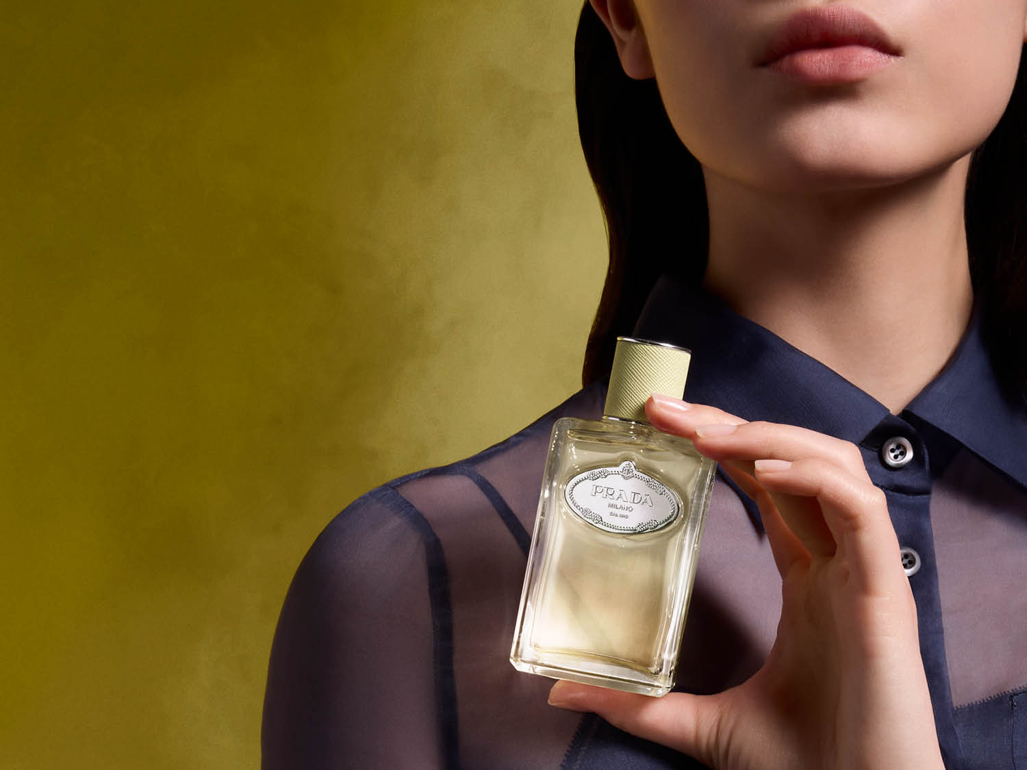 Une femme tient un flacon de parfum Prada couleur crème dans un flacon rectangulaire en verre avec un bouchon texturé vert clair.