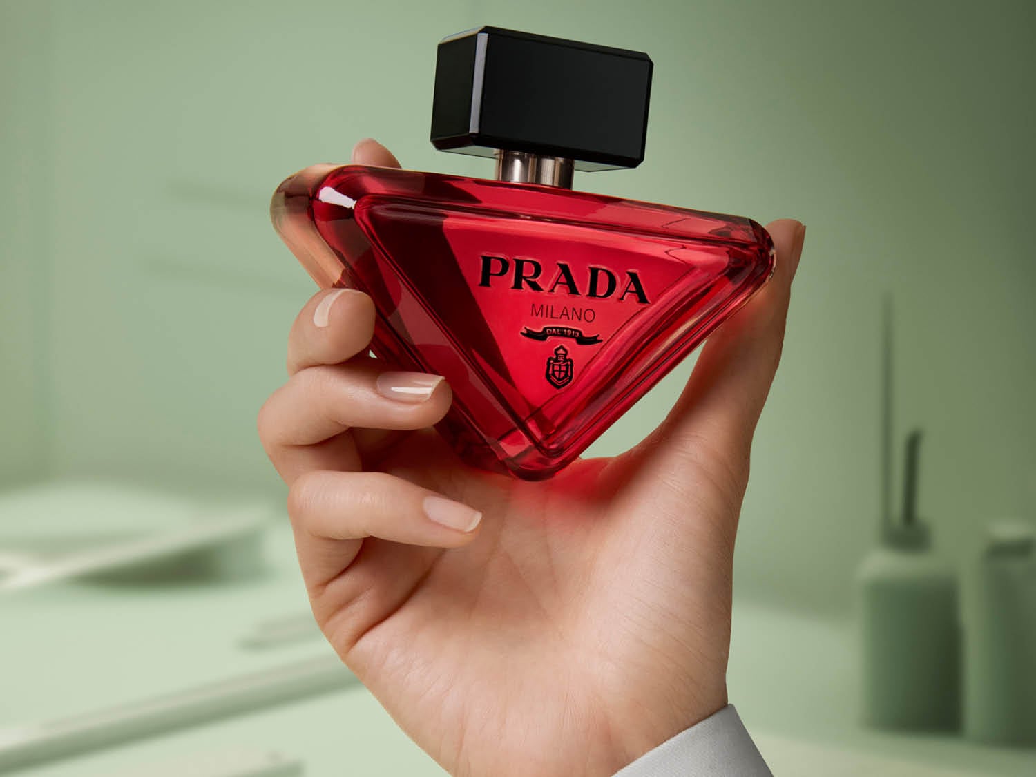 Maintenant, un parfum Prada rouge rubis est délicatement tenu dans une main, son flacon triangulaire et son bouchon noir brillant ajoutant une touche de mystère.