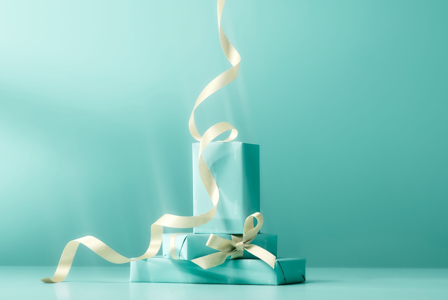 Tri tyrkysové darčekové balíčky sú na sebe poukladané a previazané zlatou stuhou, čo vytvára elegantný a slávnostný dojem.