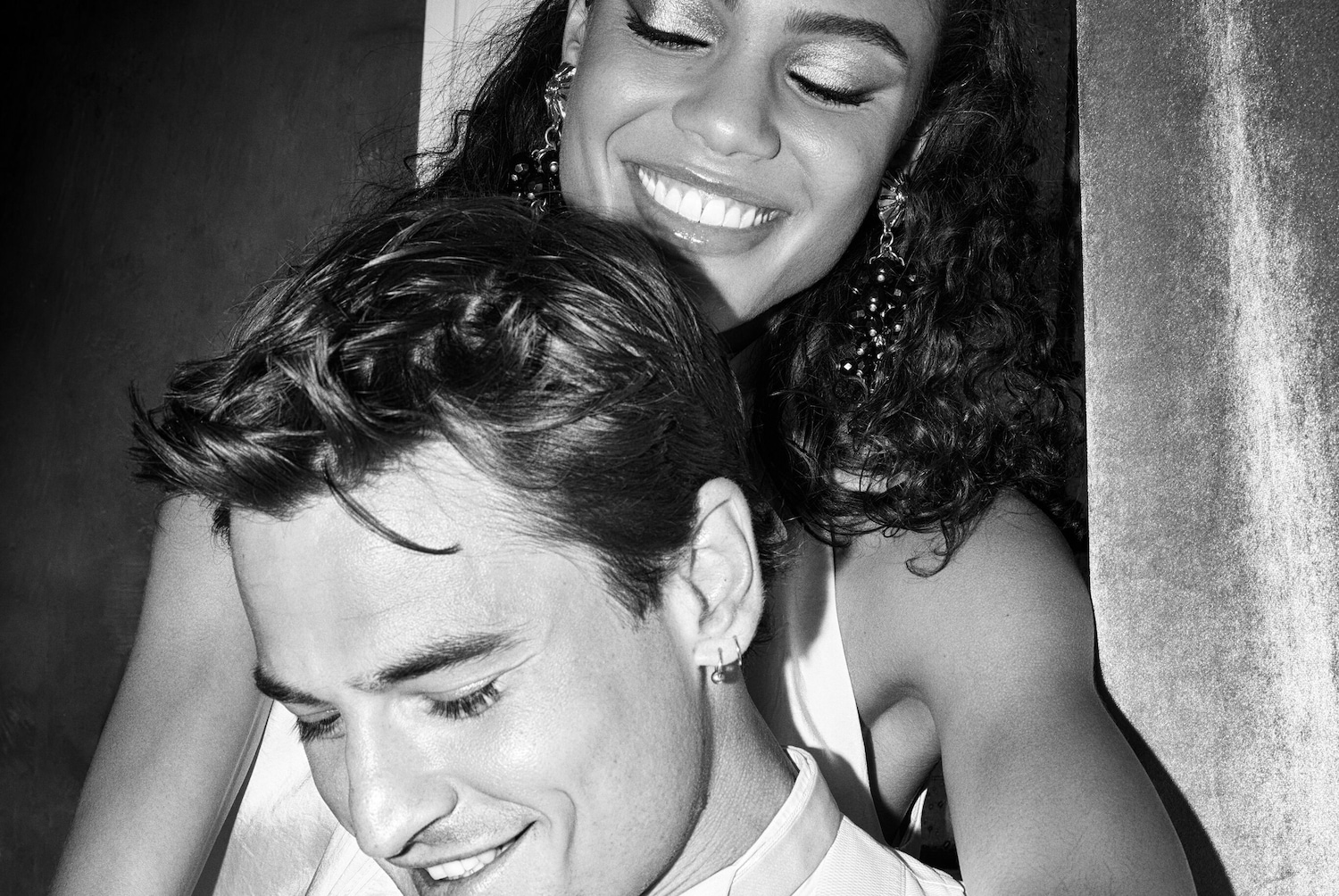 Čiernobiela fotografia usmievajúcej sa ženy s kučeravými vlasmi a náušnicami, ktorá objíma usmievajúceho sa muža.
