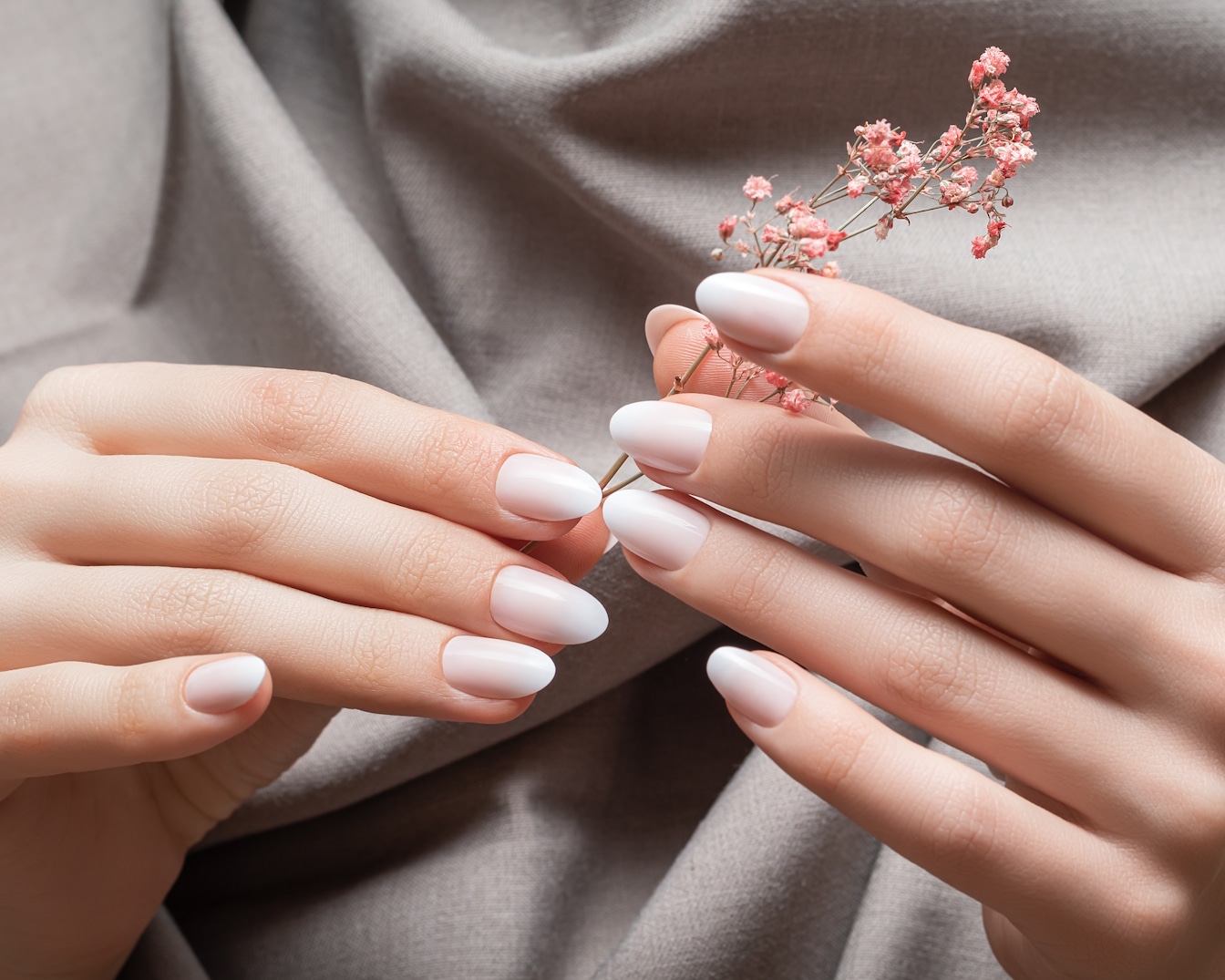 Zarte Hände präsentieren gepflegte Nägel mit einem sanften Farbverlauf von Beige zu Weiß, akzentuiert durch einen zarten rosa Blütenzweig.