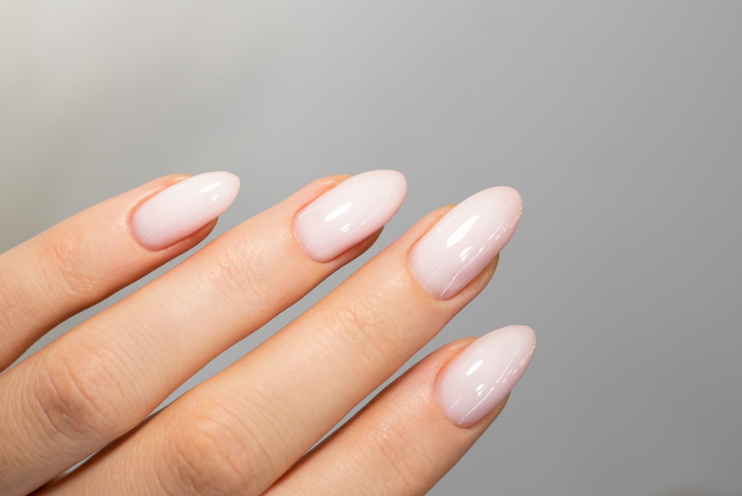 Nahaufnahme einer Hand mit mandelförmigen Nägeln, die in einem zarten, hellrosa Farbton lackiert sind und einen dezenten Glanz aufweisen.