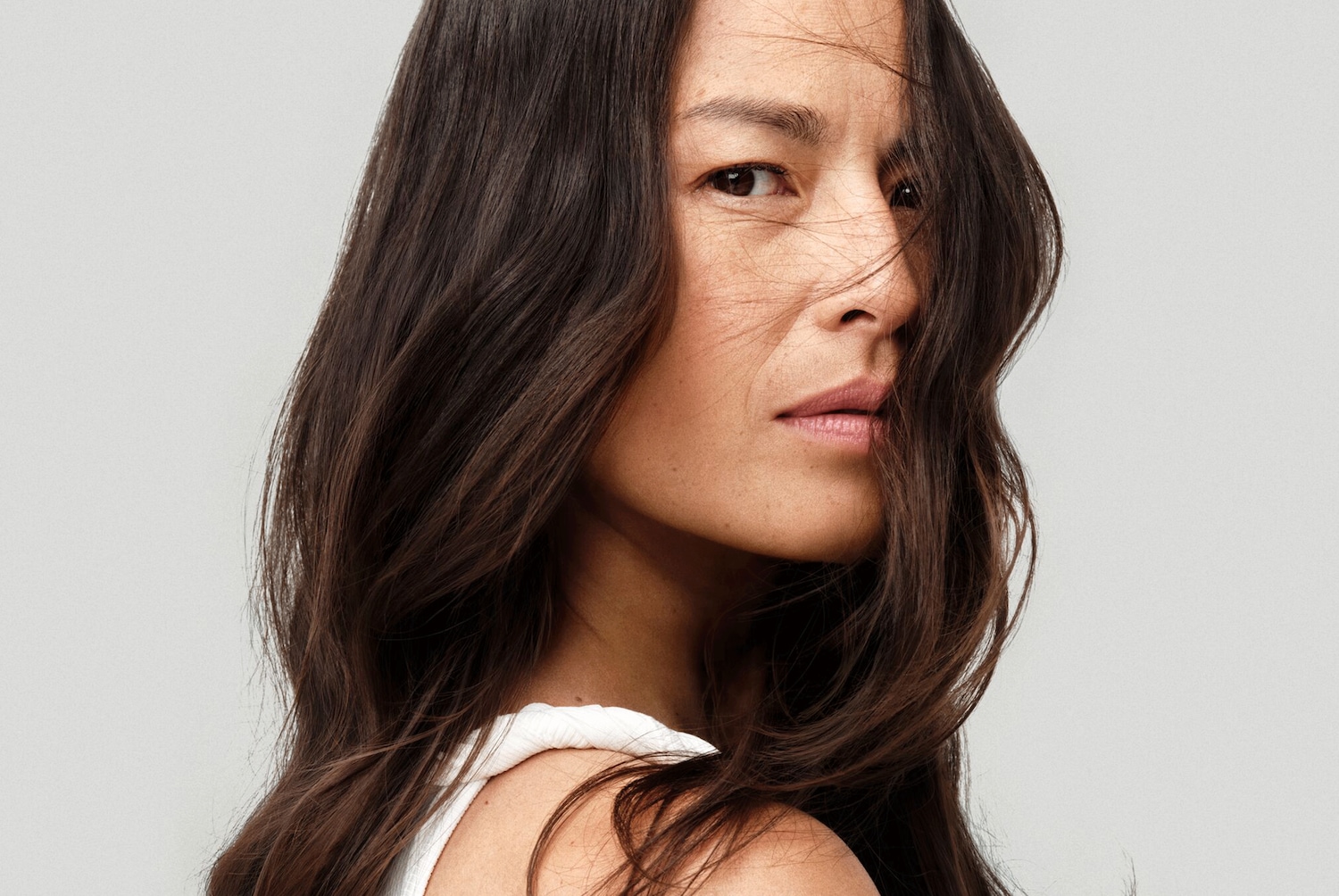 Portrait d'une femme aux longs cheveux bruns ondulés, une mèche tombant sur son visage, regardant par-dessus son épaule avec un léger sourire.