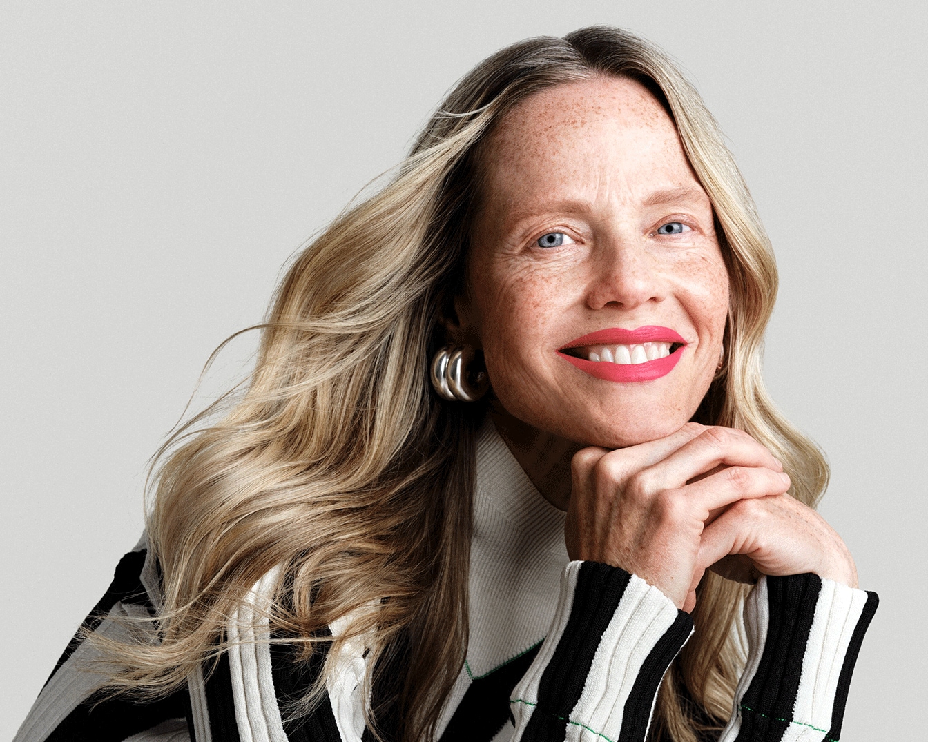 Šviesiaplaukė moteris su šypsena, ryškiai rožiniais lūpų dažais ir dryžuotu megztiniu pozuoja su sidabriniais auskarais.