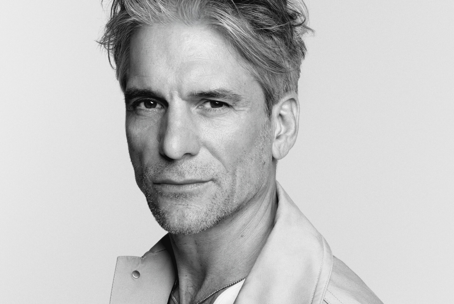 Portrait en noir et blanc d'un homme aux cheveux gris, portant une veste claire et un fin collier en chaîne.