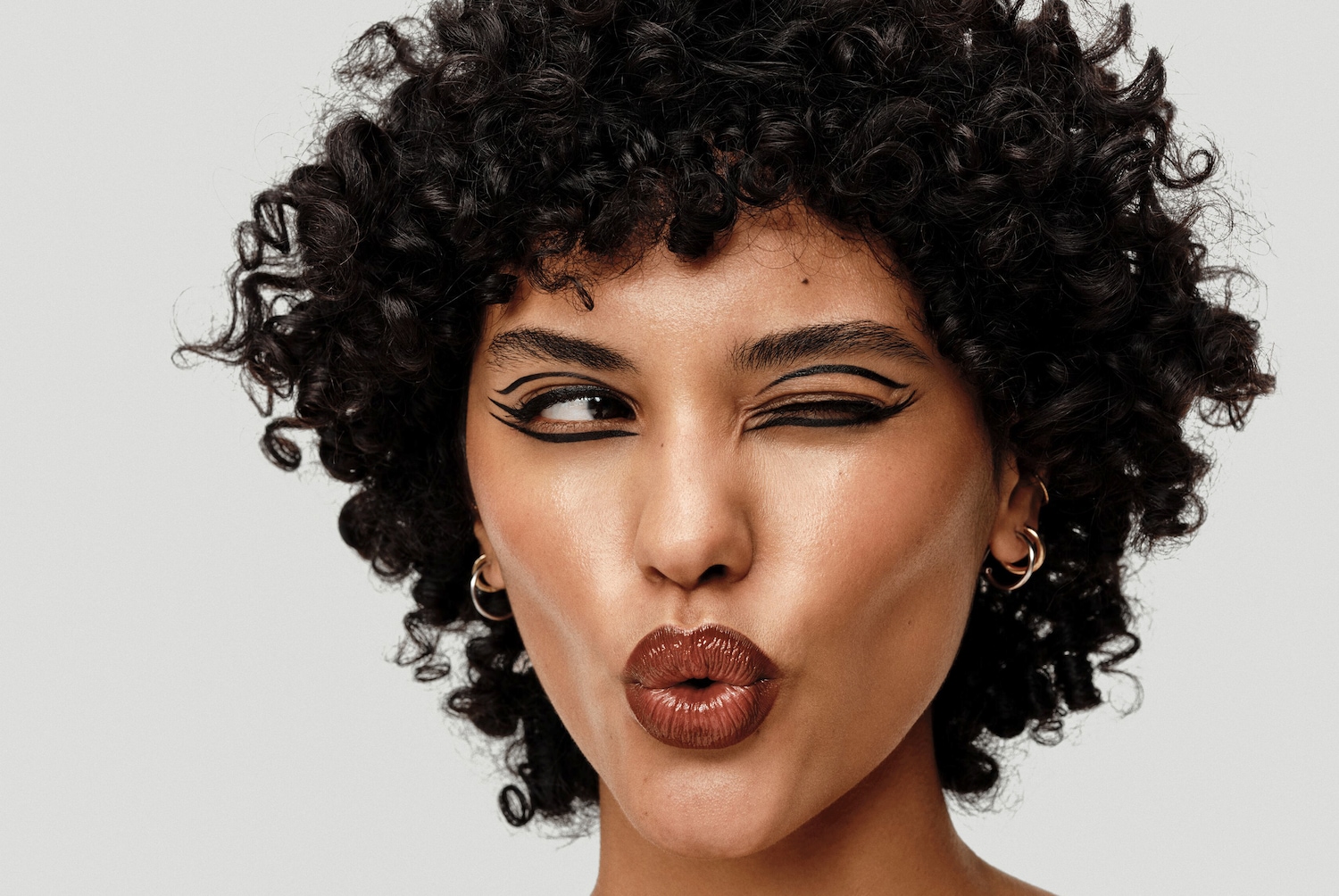 Gros plan sur un visage féminin avec un eye-liner noir graphique audacieux et des lèvres brillantes couleur bronze, le tout encadré par une chevelure bouclée et sombre.