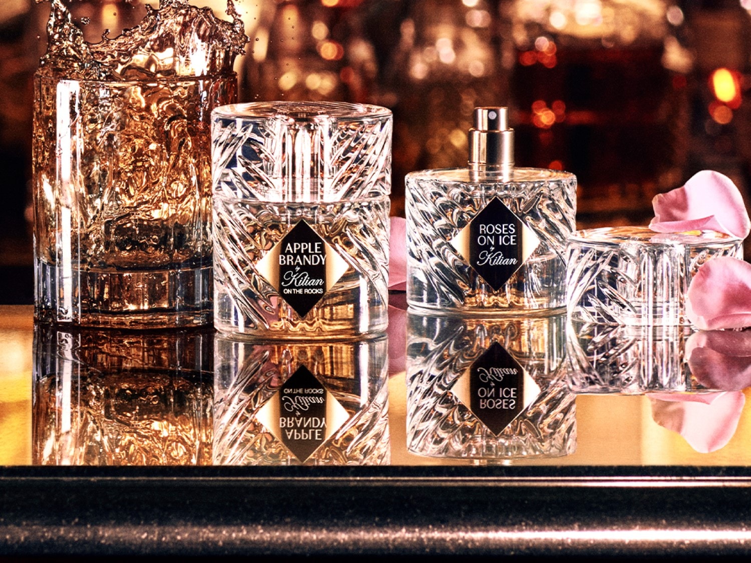 Eleganckie flakony perfum Kilian „Apple Brandy on the Rocks” i „Roses on Ice” z przezroczystego szkła o fasetowanej strukturze, stojące na błyszczącej powierzchni obok kieliszka z trunkiem i płatków róż.