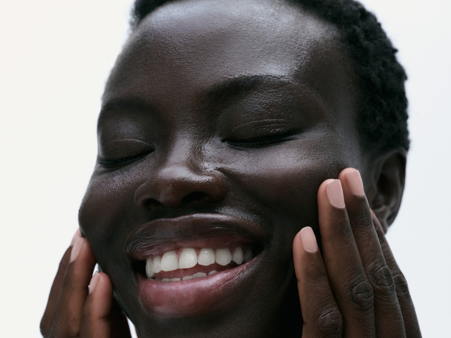 Une femme noire souriante, les yeux fermés, touche sa peau brillante, mettant en valeur une texture lisse et hydratée.
