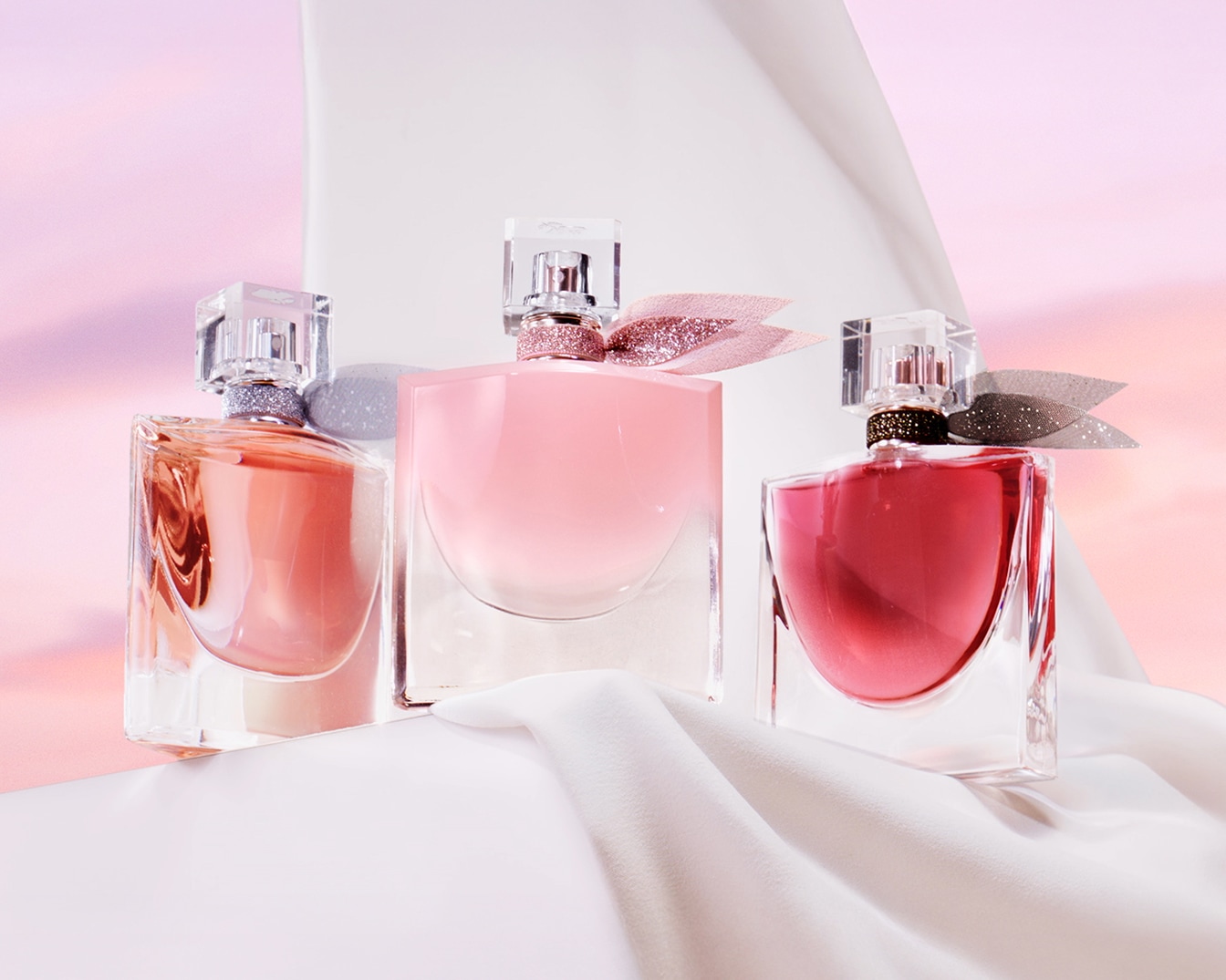 Découvrez un trio de parfums La Vie Est Belle dans des flacons en verre transparent aux teintes rose, pêche et rouge, ornés de nœuds scintillants, reposant sur un tissu blanc soyeux.