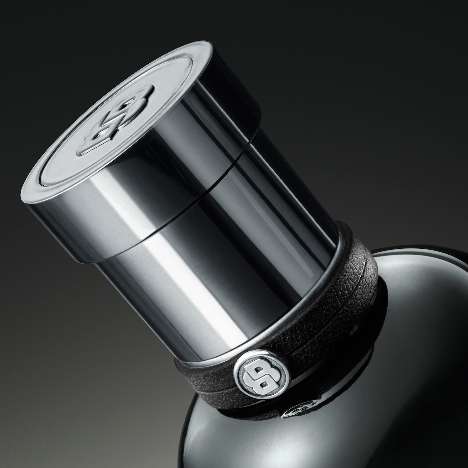 Primo piano di un flacone di profumo nero lucido con tappo cilindrico argentato e cinturino in pelle con logo.