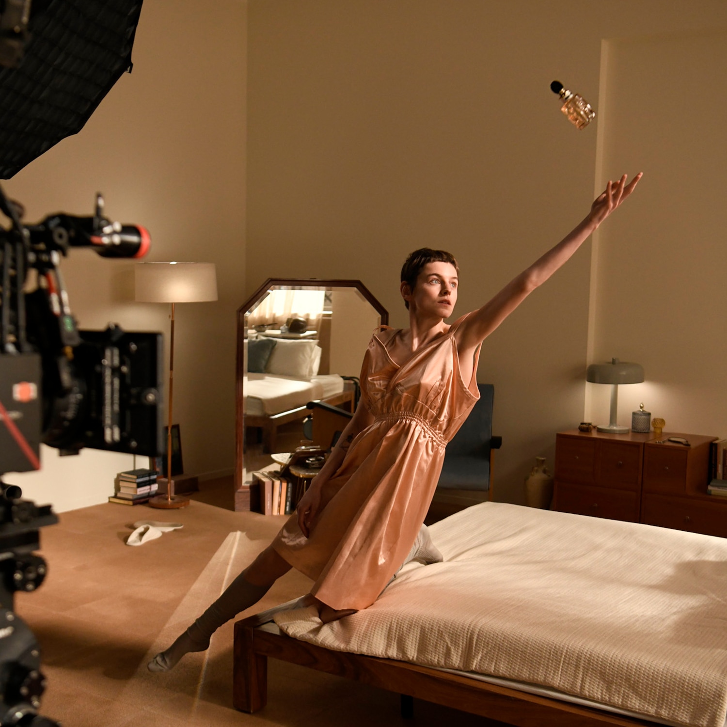 Dans une chambre chaleureuse, une femme en robe attrape un flacon de parfum en l'air, capturée par une caméra professionnelle.