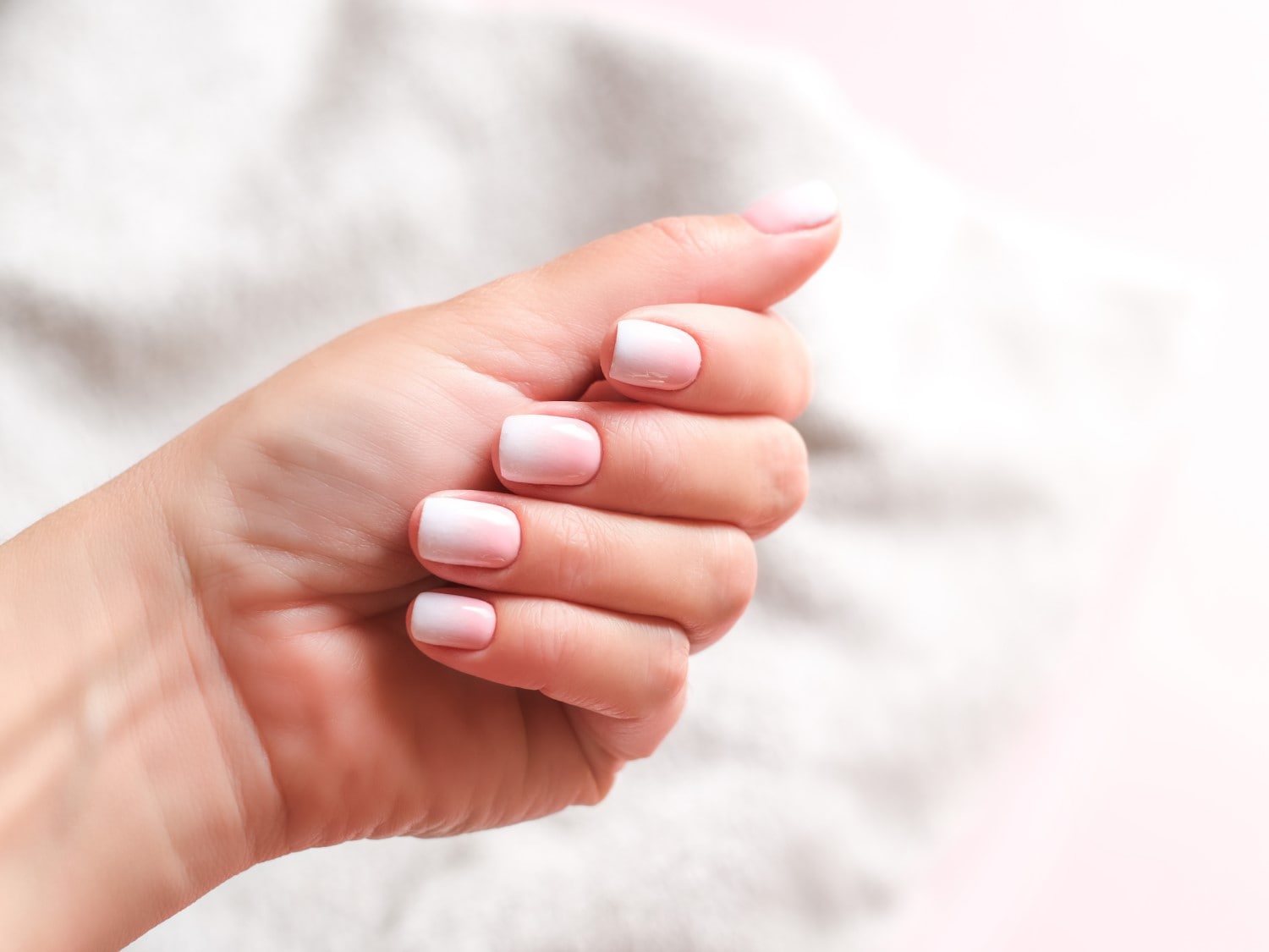 Nahaufnahme einer Hand mit Ombre-Maniküre, die von einem zarten Rosa zu einem sauberen Weiß übergeht und einen eleganten und gepflegten Look erzeugt.