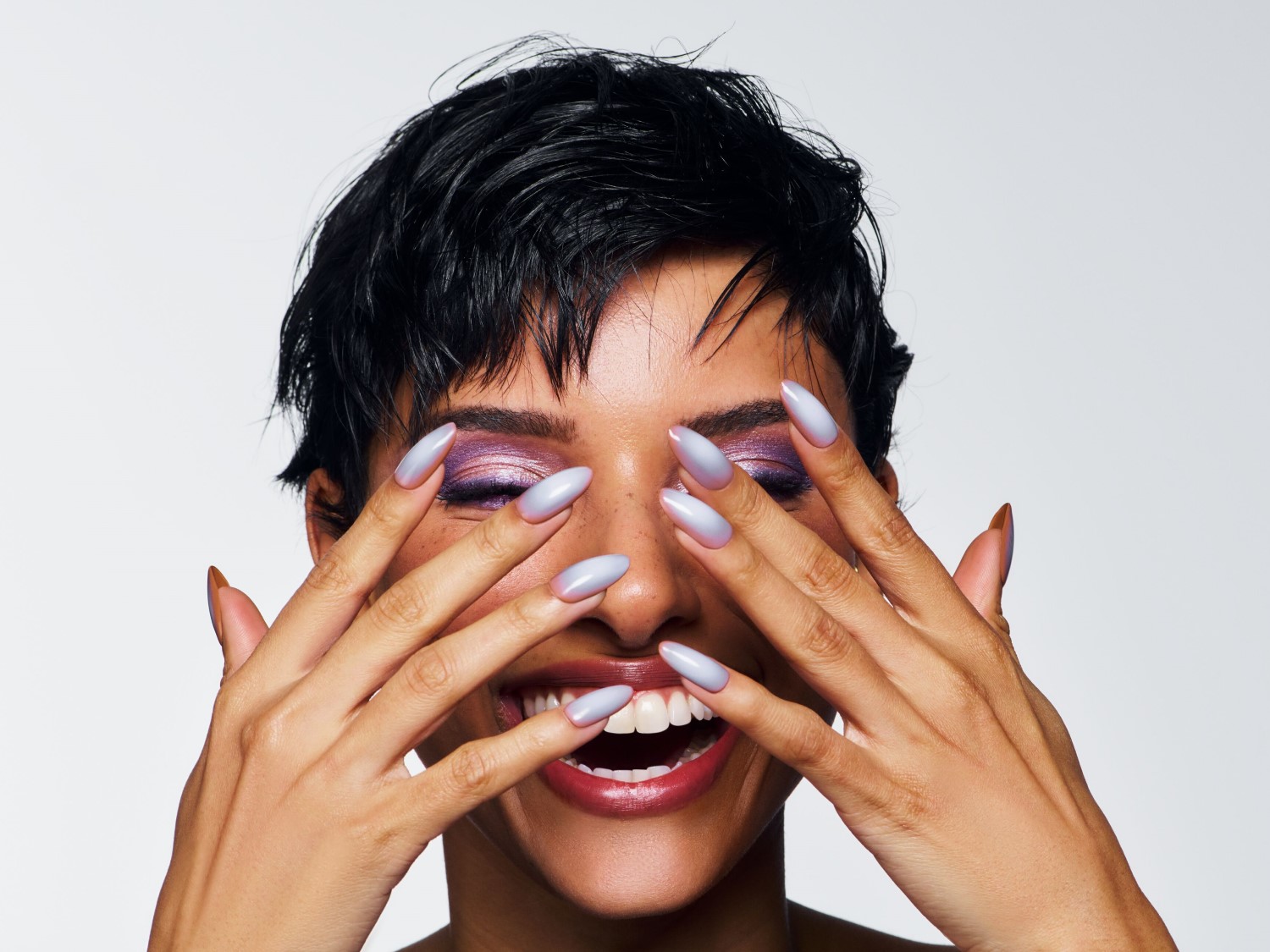 Eine strahlende Frau mit lila Lidschatten und hellblauen Nägeln bedeckt ihr Gesicht mit den Händen und lächelt.