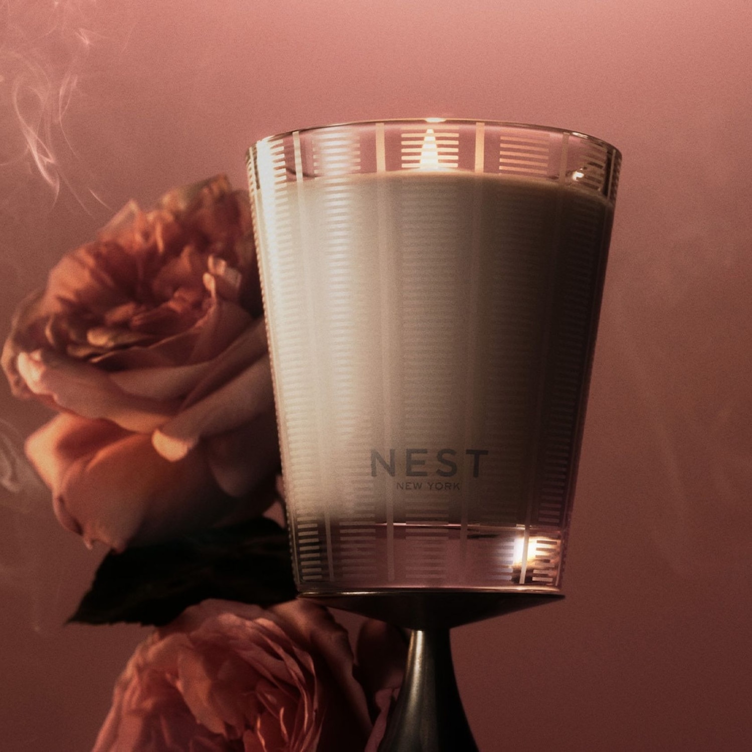A Nest New York gyertya egy elegáns üvegpohárban, rózsaszín virágok között, lágy füsttel körülvéve.