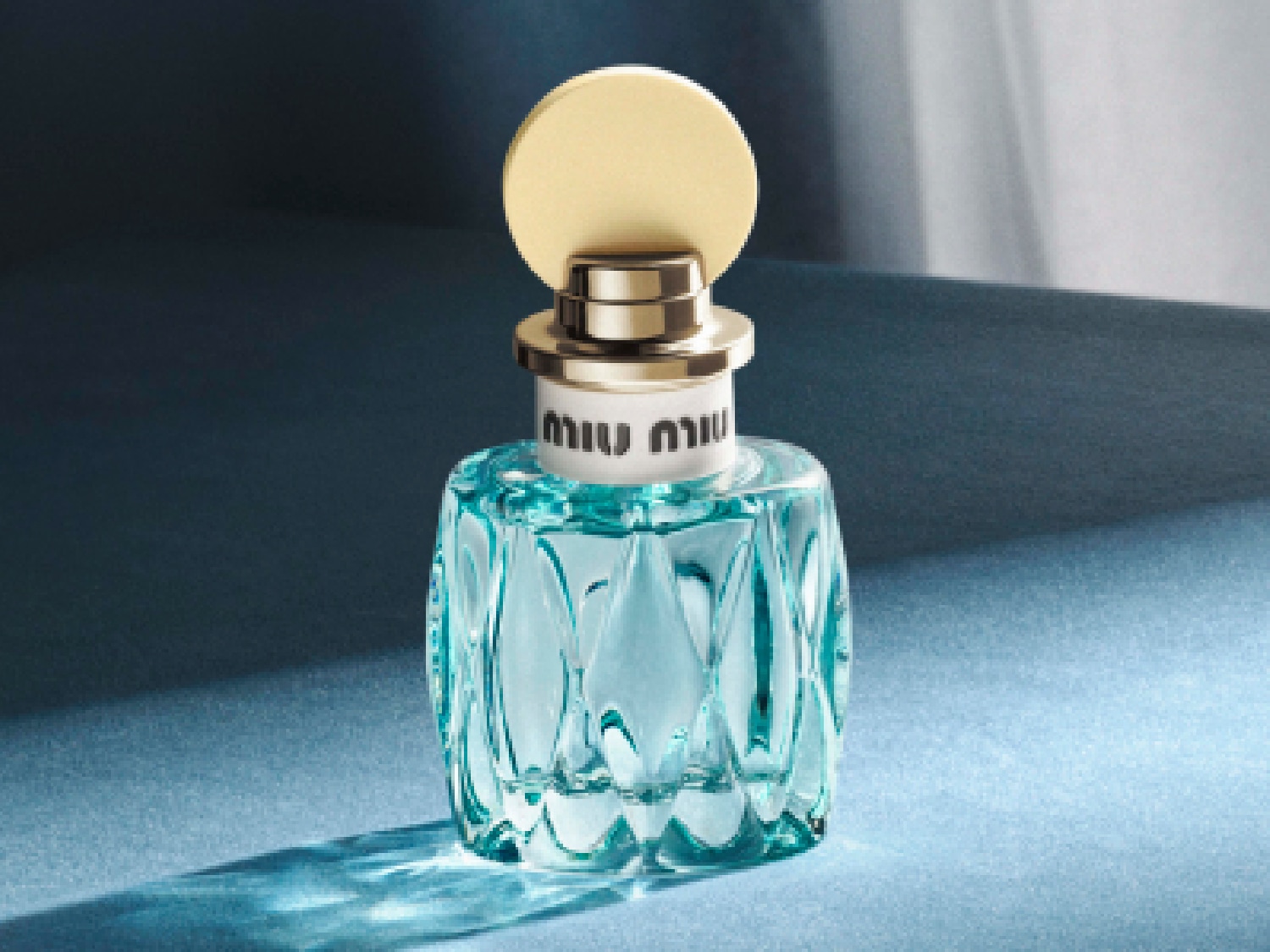 Le flacon de parfum Miu Miu bleu clair aux facettes complexes est coiffé d'un bouchon doré, évoquant une élégance rafraîchissante.