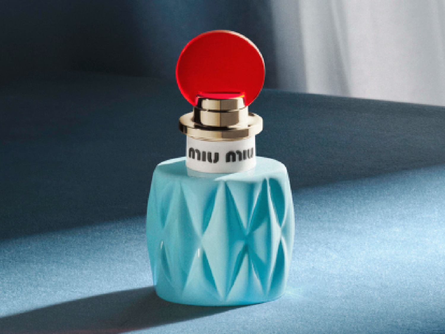 Découvrez le parfum Miu Miu, présenté dans un flacon bleu turquoise aux motifs géométriques, surmonté d'un bouchon rouge vif et doré.