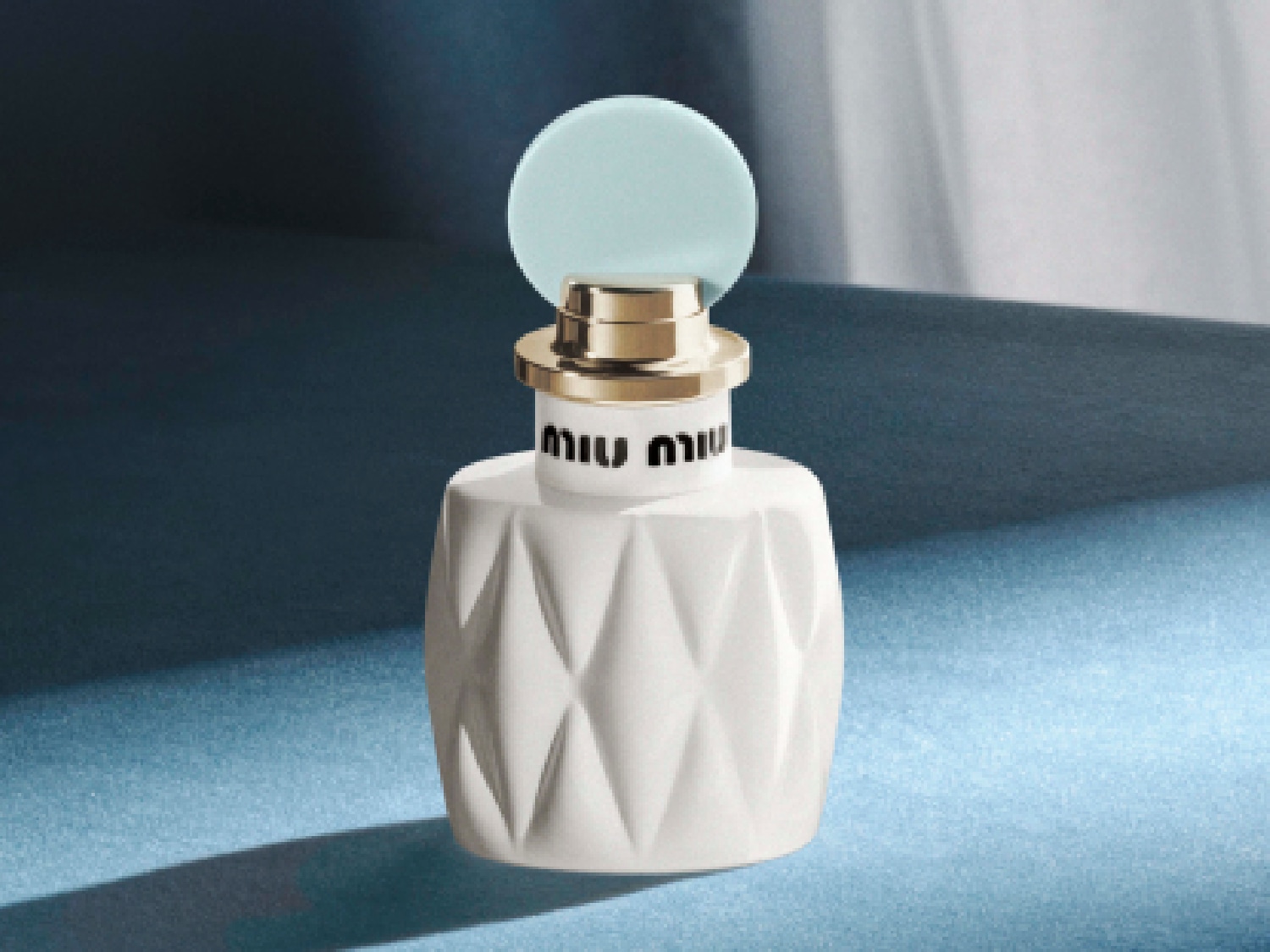 Découvrez le parfum Miu Miu, présenté dans un flacon blanc aux motifs géométriques, surmonté d'un bouchon bleu clair et doré, évoquant luxe et modernité.