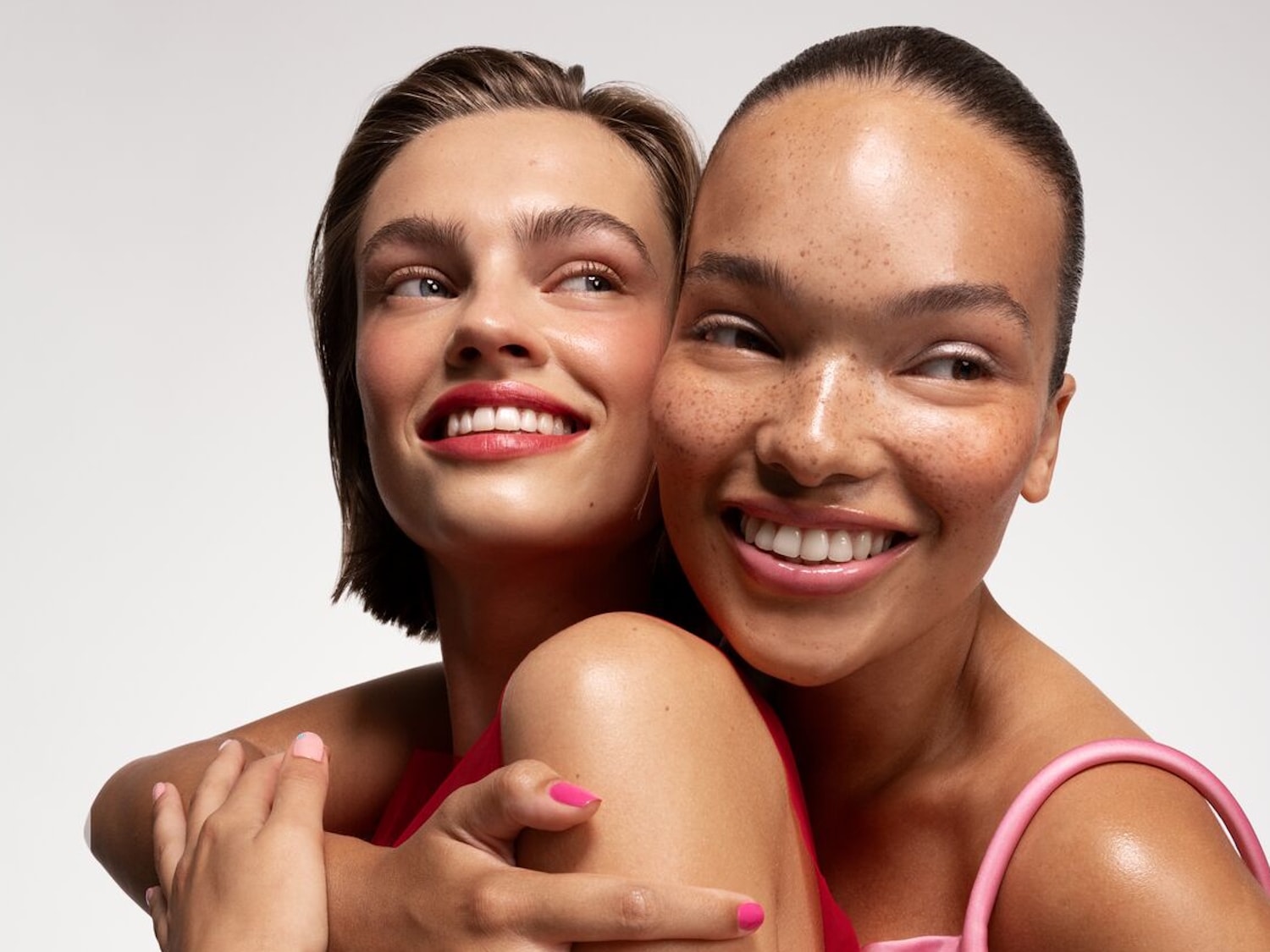 Zwei strahlende Frauen umarmen sich, ihre Haut schimmert mit einem Hauch von Rouge und Lipgloss, der ihre natürliche Schönheit hervorhebt.