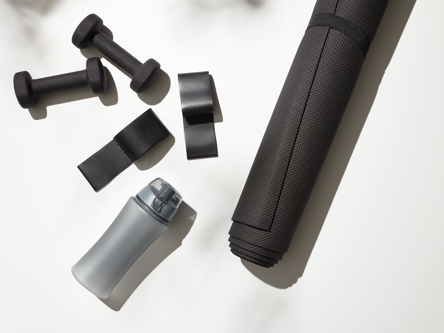 Draufsicht auf eine graue Wasserflasche, zwei schwarze Hanteln, drei schwarze Widerstandsbänder und eine zusammengerollte schwarze Yogamatte auf einem weißen Hintergrund.